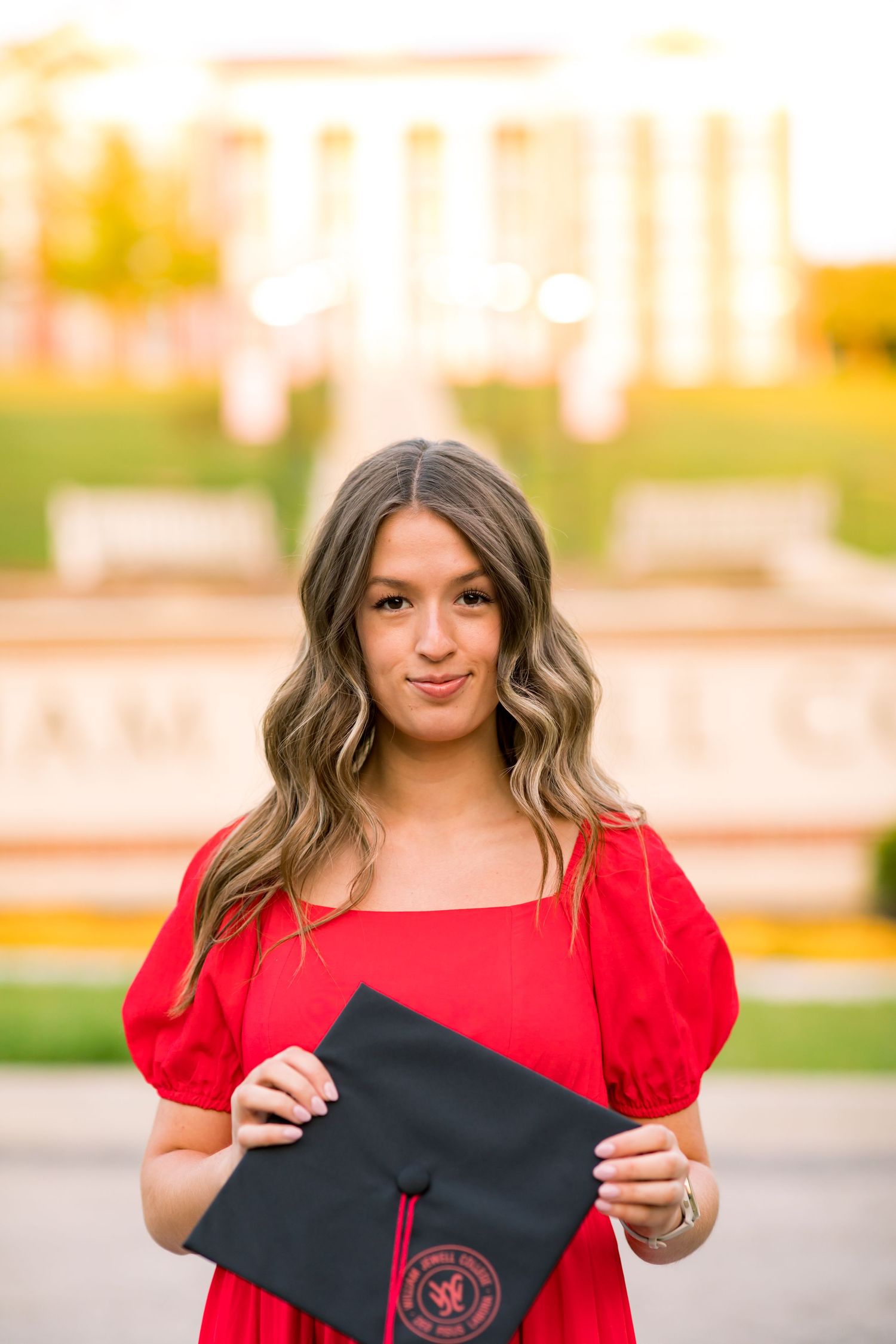 A graduate in a red dress holds a black graduation cap with red emblem outdoors during golden hour.
