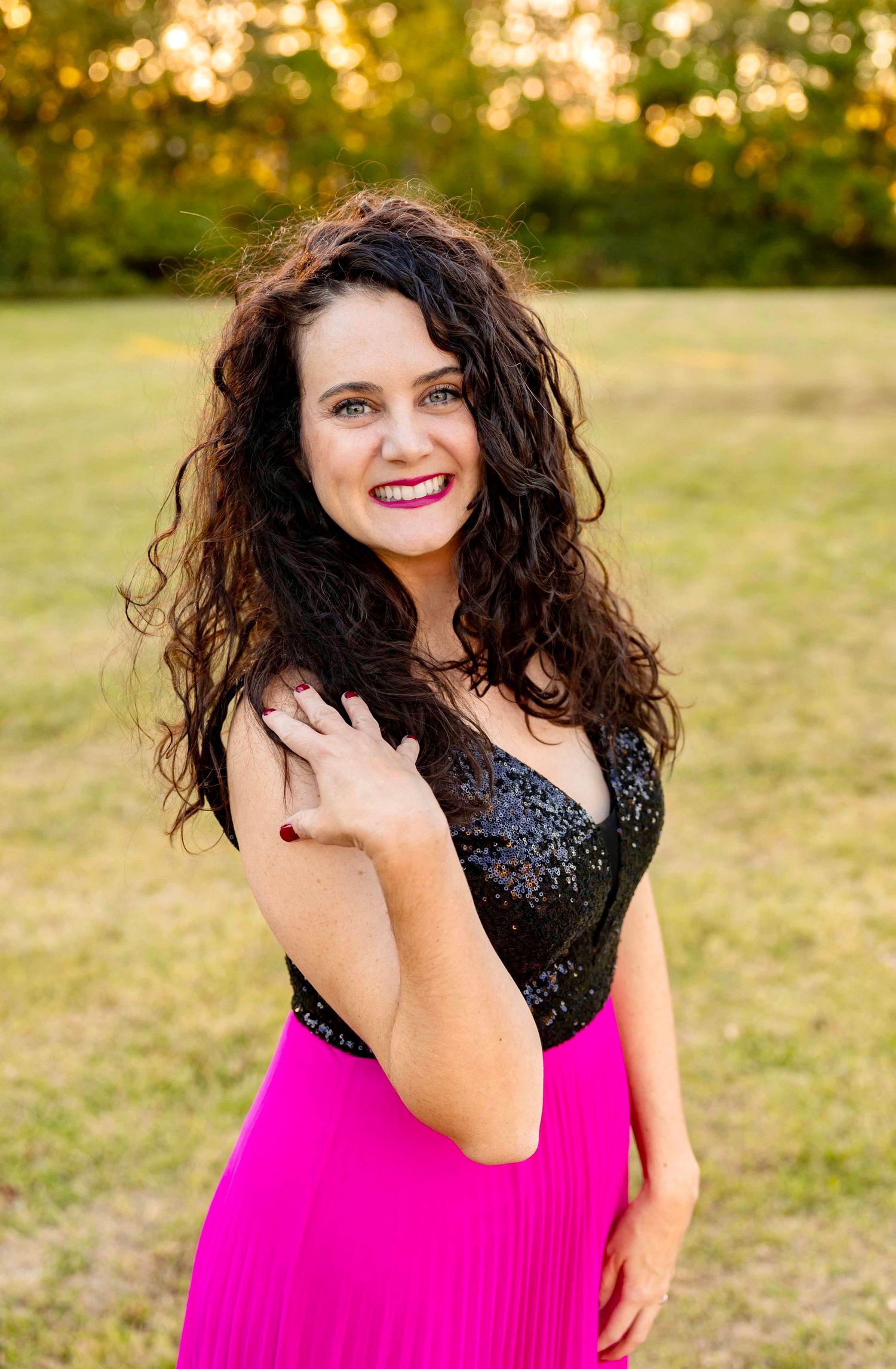 A woman in a black sequined top and bright hot pink skirt smiles outdoors during golden hour.
