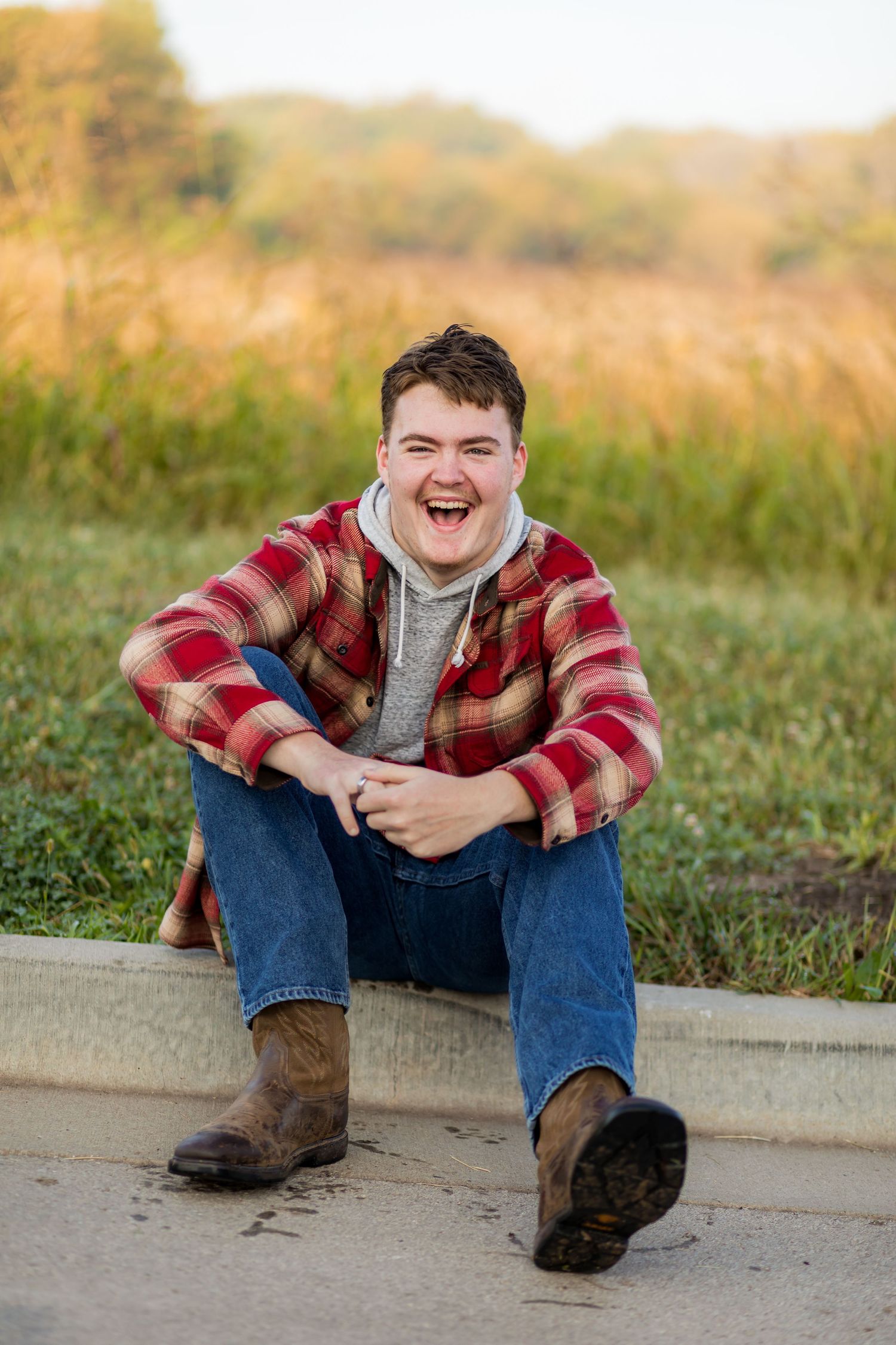 Someone sits casually on a curb wearing a plaid jacket, jeans and boots against an autumn field background.