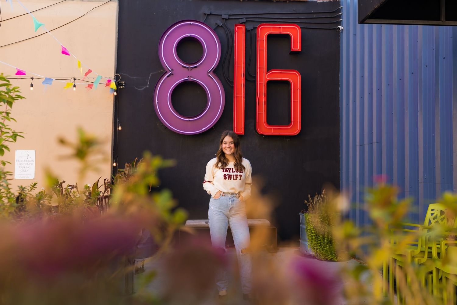 A person poses against a dark wall with bright neon numbers '816' while flowers bloom in the foreground.