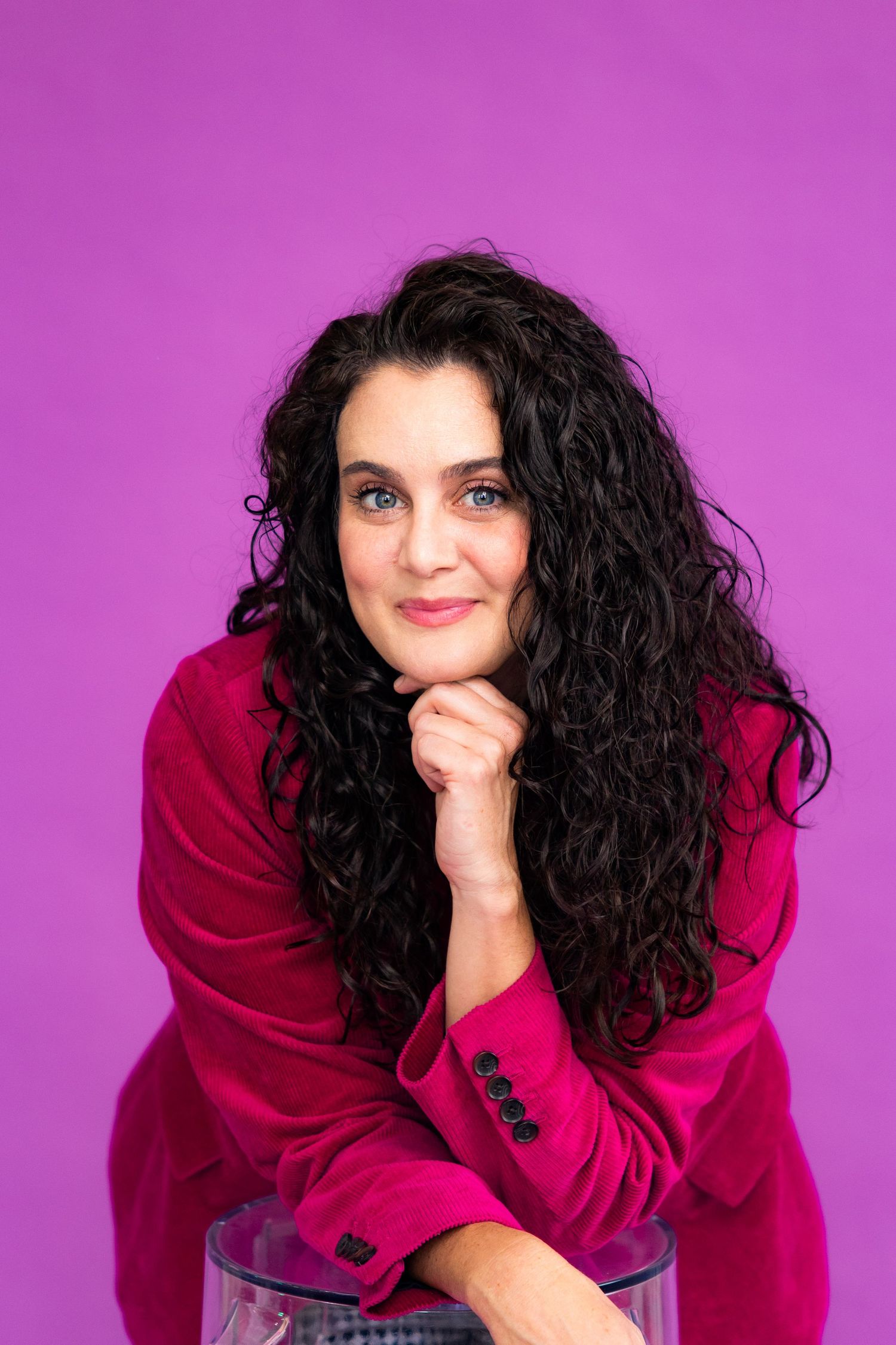 Woman in red blazer with dark curly hair poses against bright magenta backdrop in studio portrait.