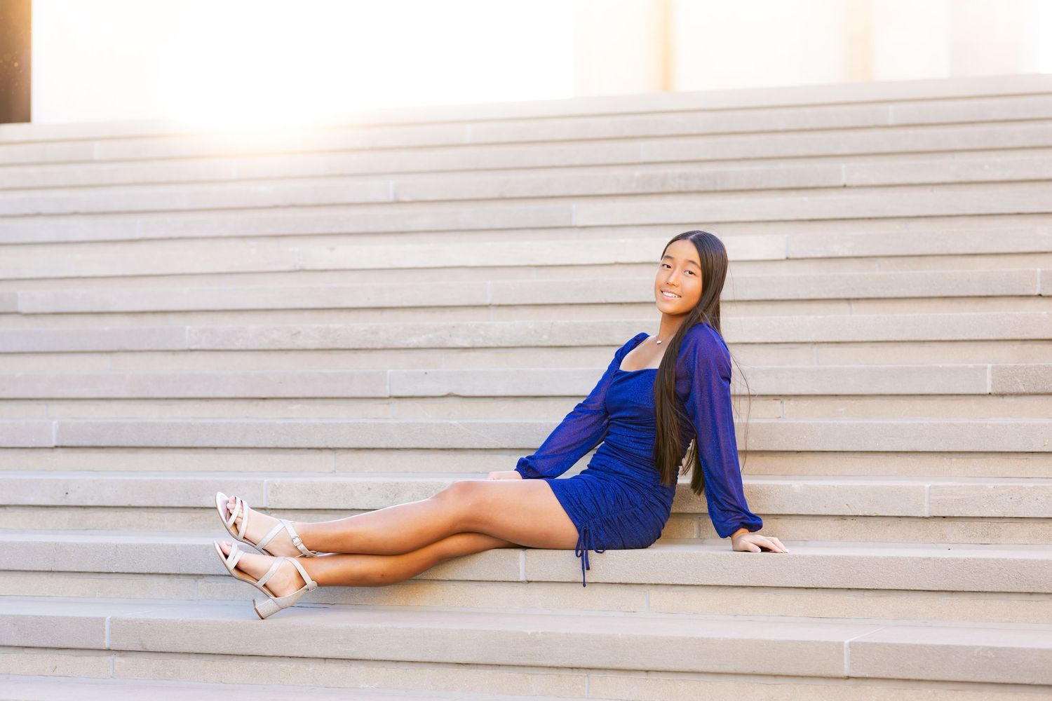 Person in a bright blue dress sitting comfortably on concrete steps while basking in sunlight.