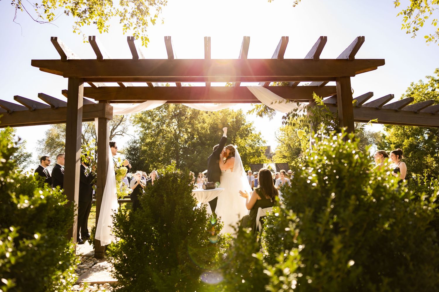 A romantic wedding ceremony under a wooden pergola backlit by golden sunlight in a lush garden setting.