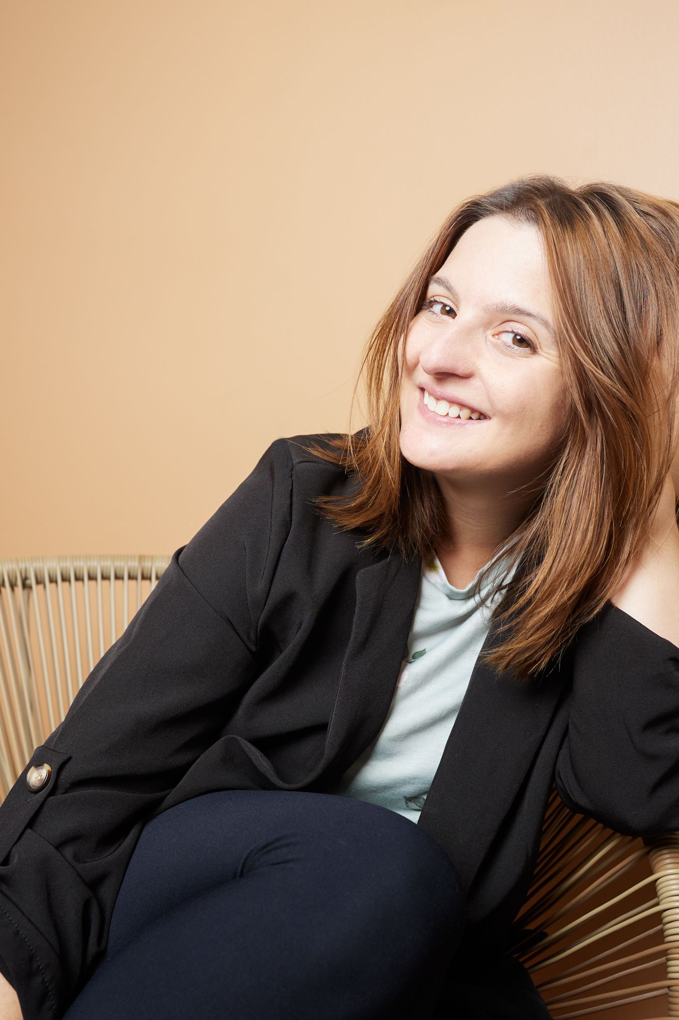 Professional portrait of woman in black blazer and light blue top lounging on modern chair against beige background.
