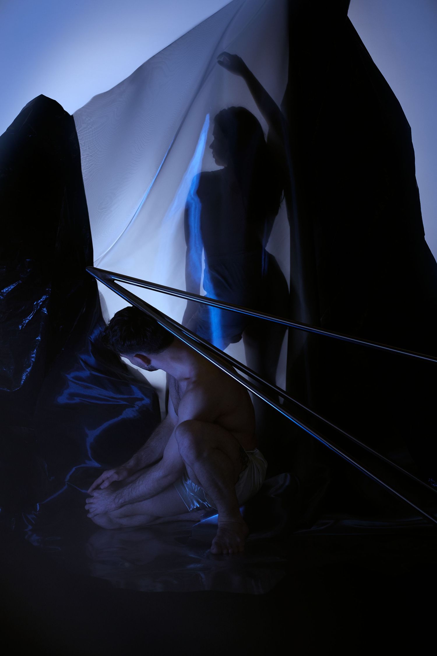 Artistic silhouette in blue lighting behind translucent fabric creates dramatic shadows and shapes in dark space.