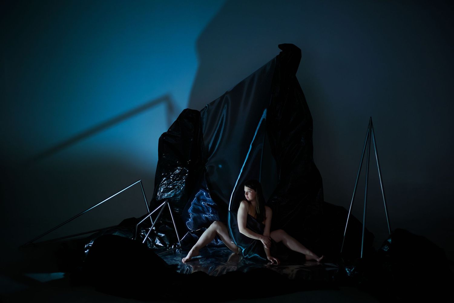 Dark artistic performance piece in moody blue lighting with draped fabric and angular geometric shapes creating shadows.