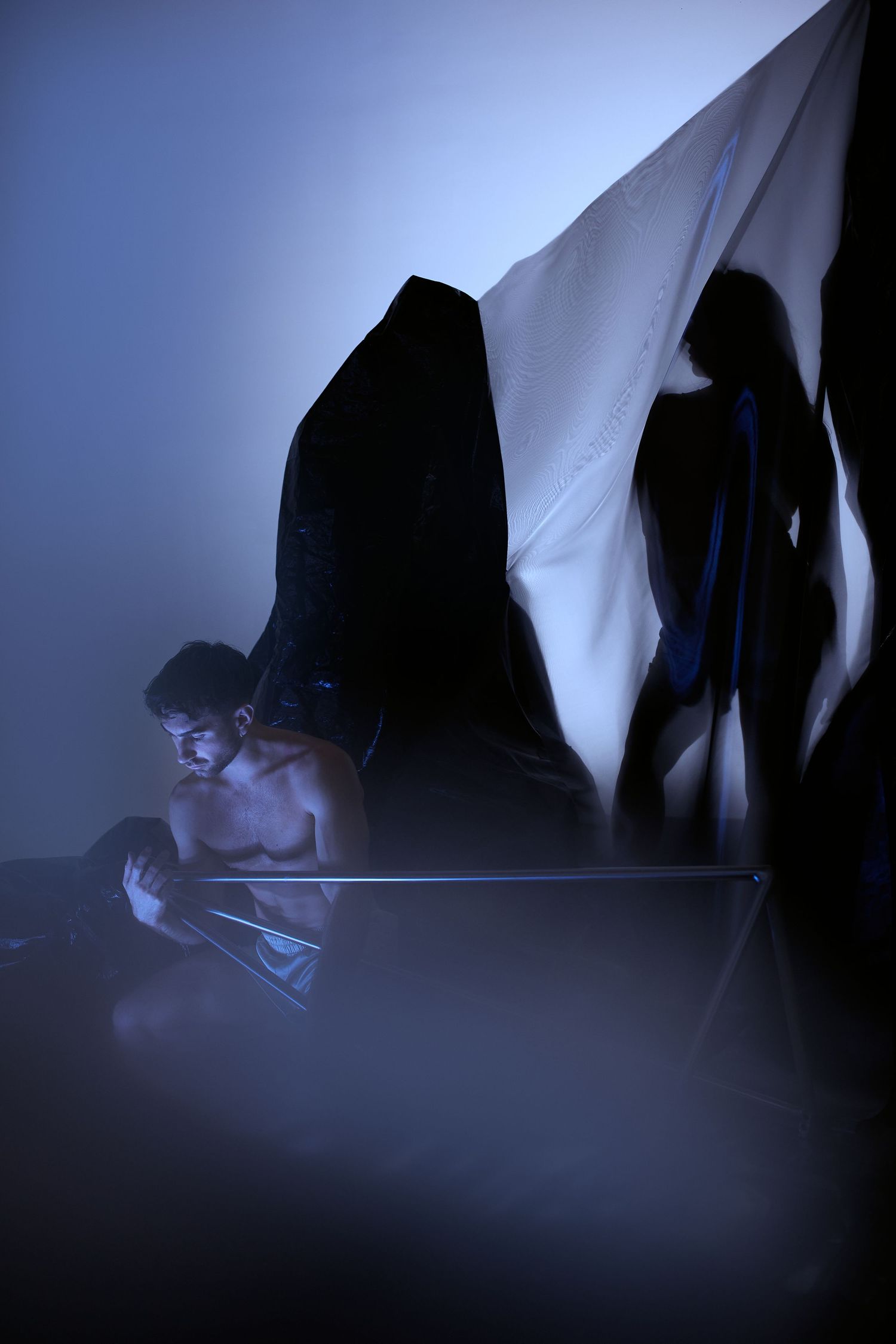 Dramatic silhouette scene with person reading in blue misty light while shadows loom on white fabric backdrop.