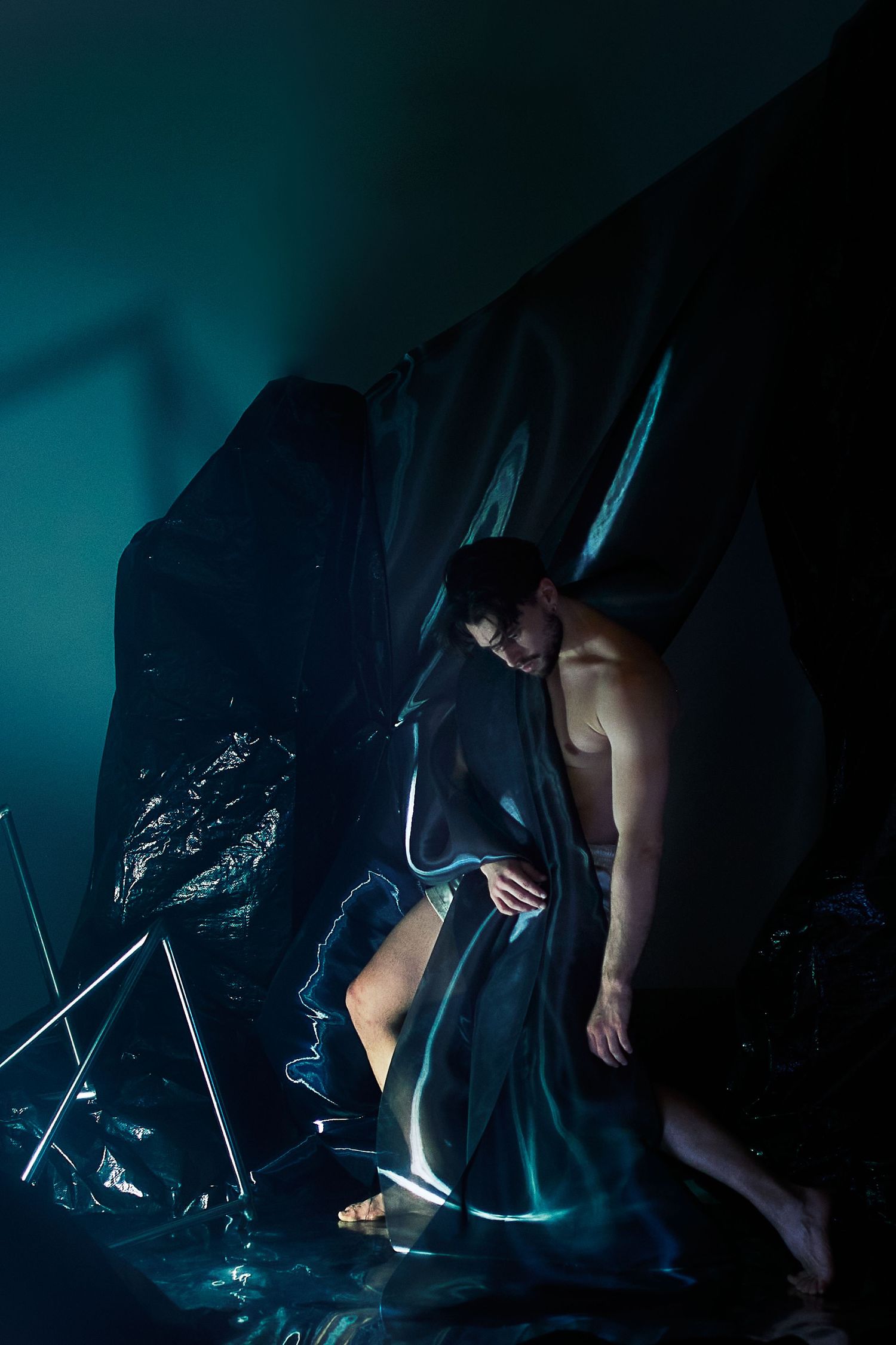 A dramatic artistic photo showing a person draped in dark fabric against a moody teal-colored backdrop.