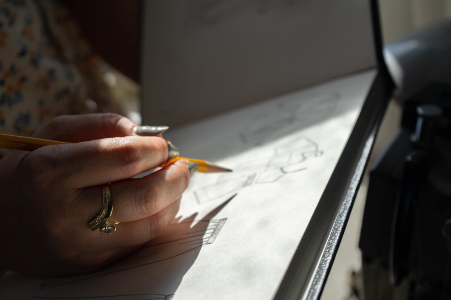 A hand holding a pencil writes on paper near a sunlit window casting soft shadows.