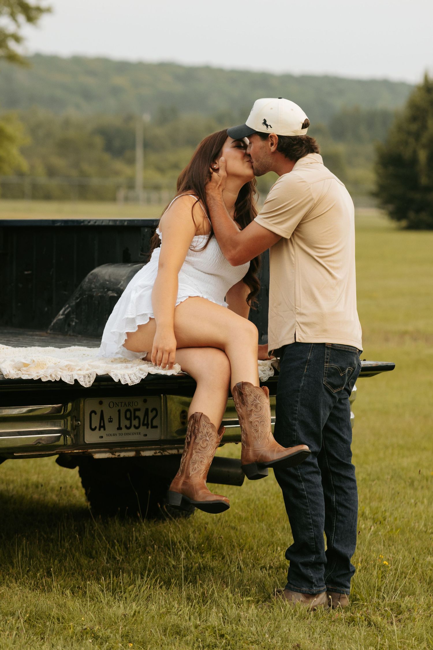 Romantic country scene with couple in cowboy boots sitting on pickup truck tailgate in green field at sunset.