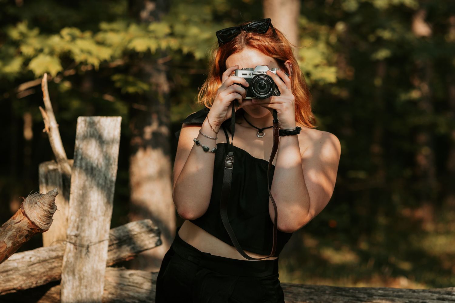 A photographer in black attire takes a photo with a vintage camera in a sunlit outdoor setting.