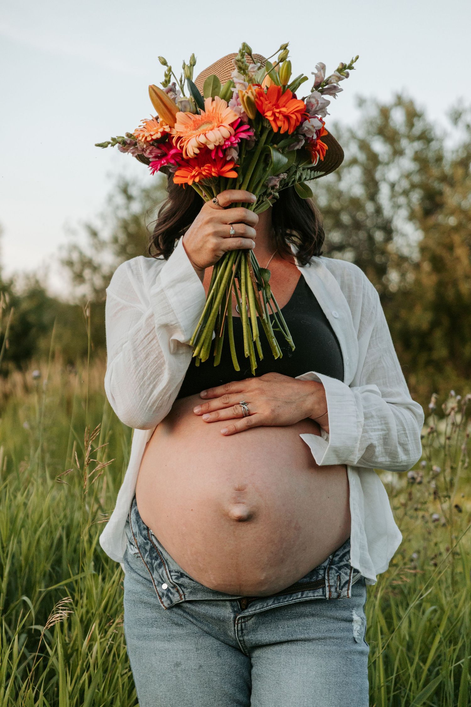 A bouquet of vibrant orange and coral flowers held against a pregnant belly in a sunlit meadow at golden hour.