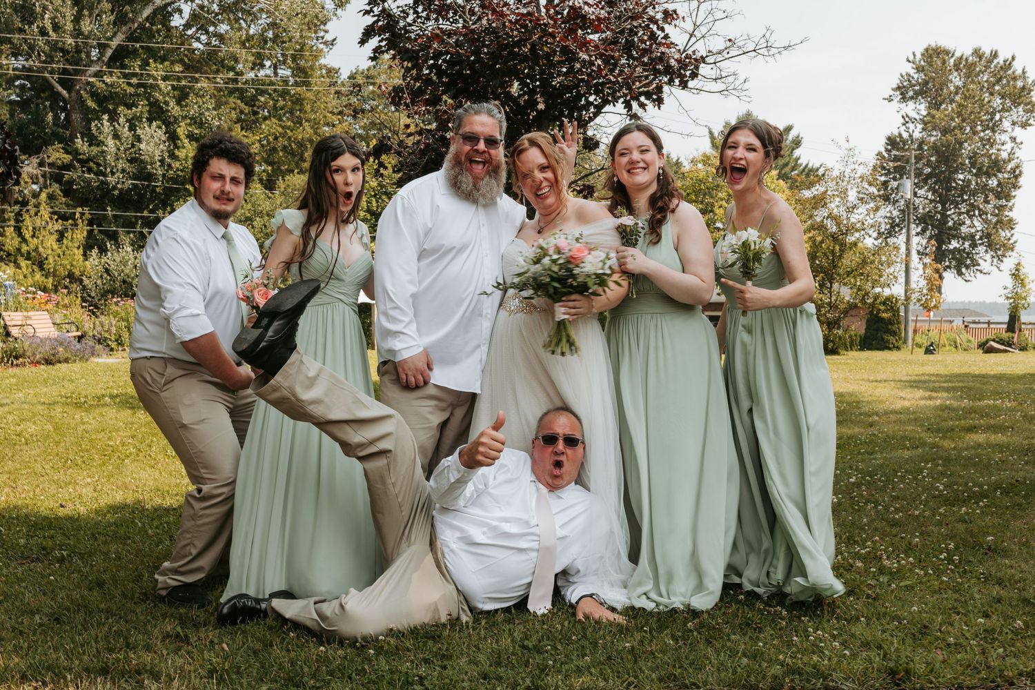Wedding party wearing sage green dresses and white shirts poses playfully on grassy lawn at outdoor celebration.