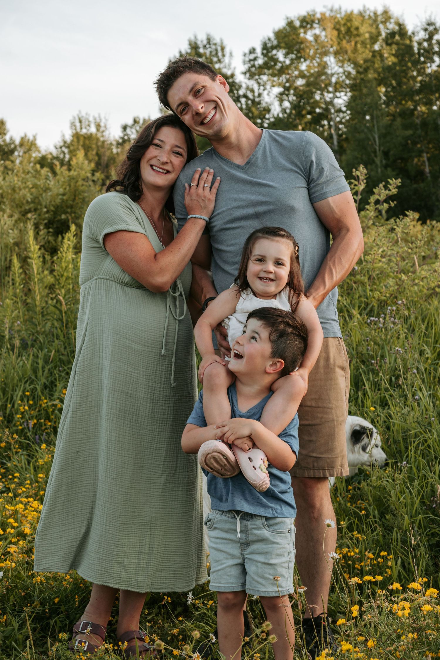 A family stands together laughing and smiling in a meadow with yellow wildflowers and trees in the background.