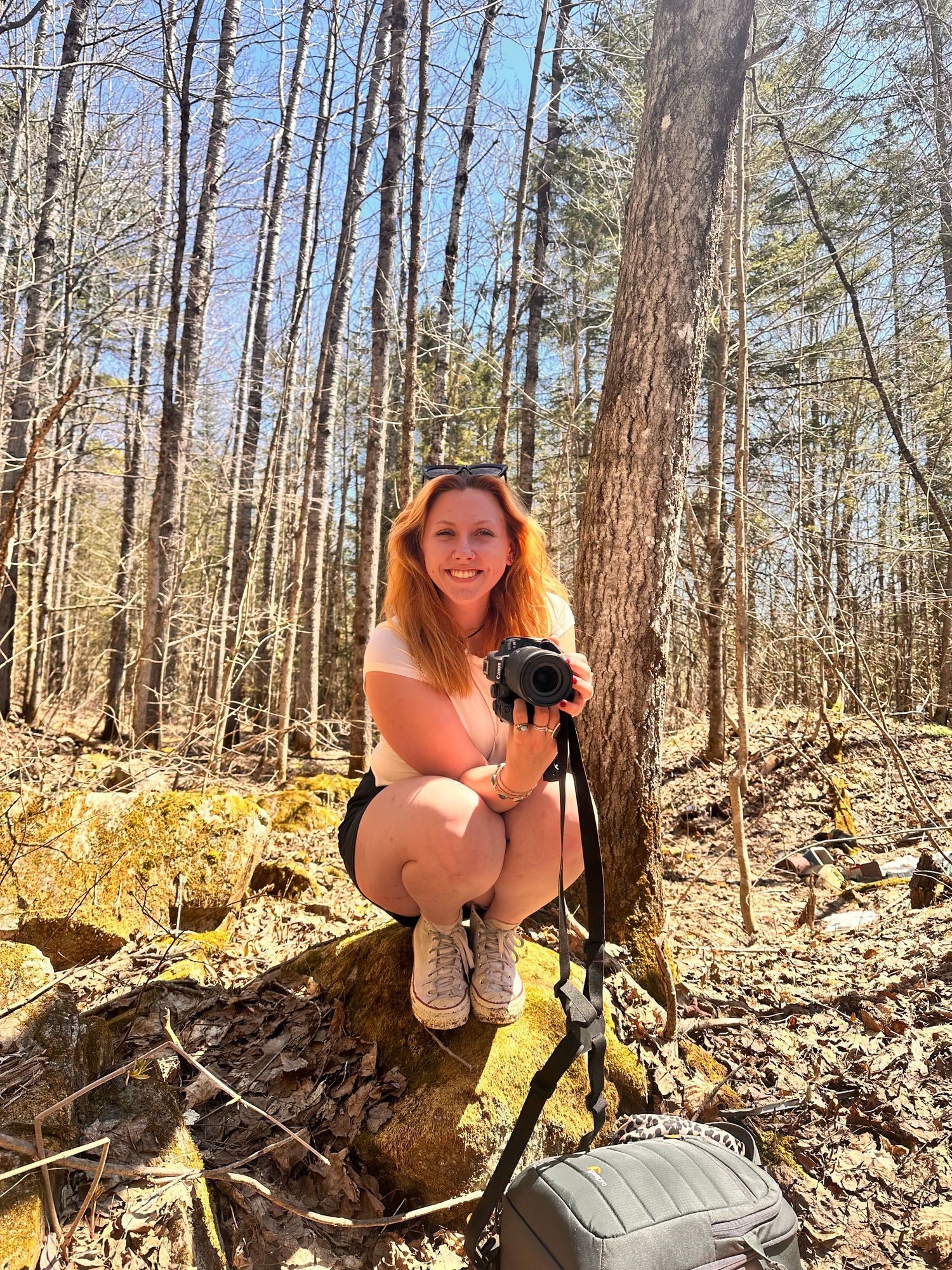 A hiker crouches on a mossy rock in a sunlit forest while holding camera equipment and wearing hiking gear.