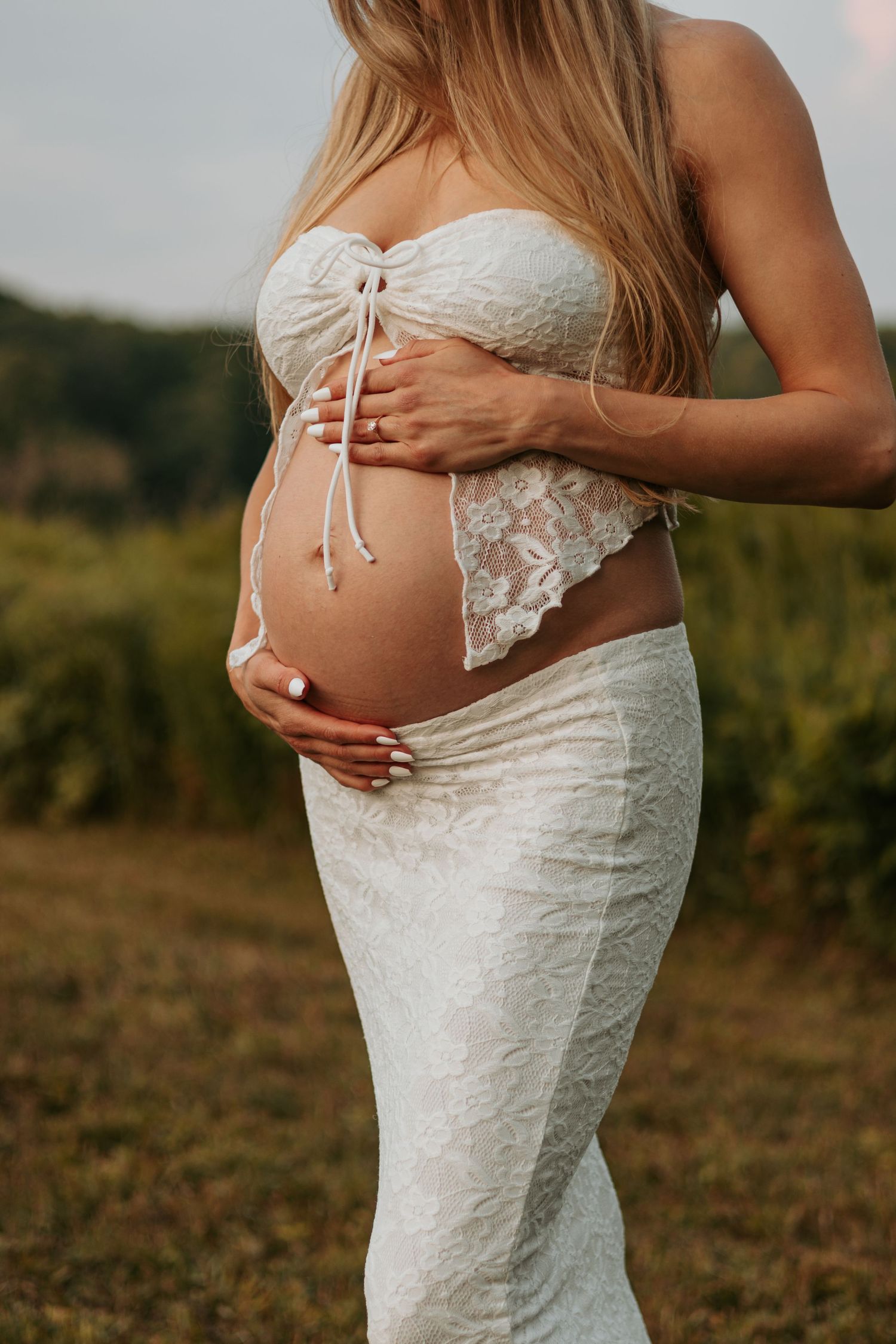 Closeup of pregnant belly in white lace maternity dress during golden hour photoshoot outdoors.
