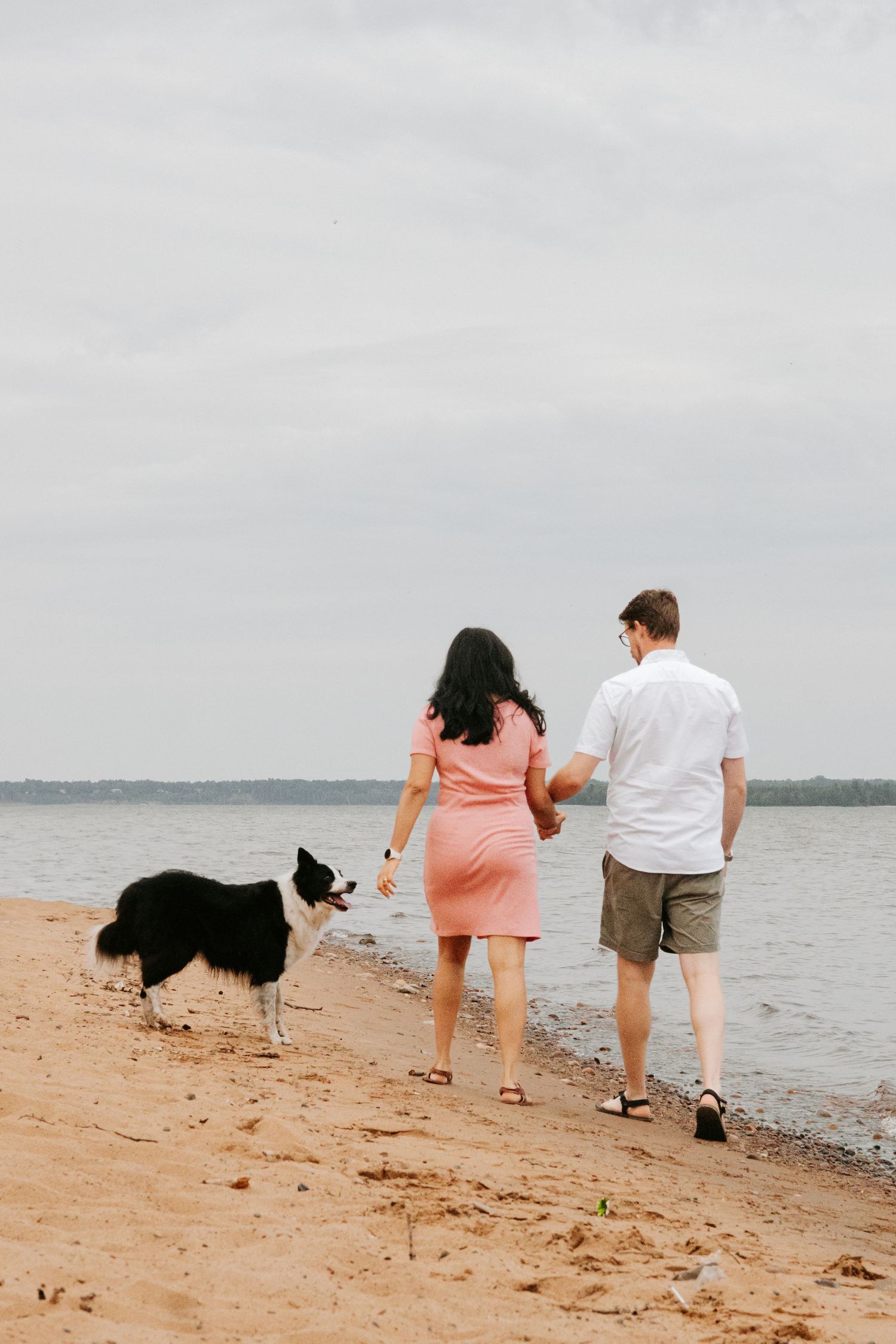 Couple walking with their black Border Collie dog along a sandy beach shoreline on an overcast day.