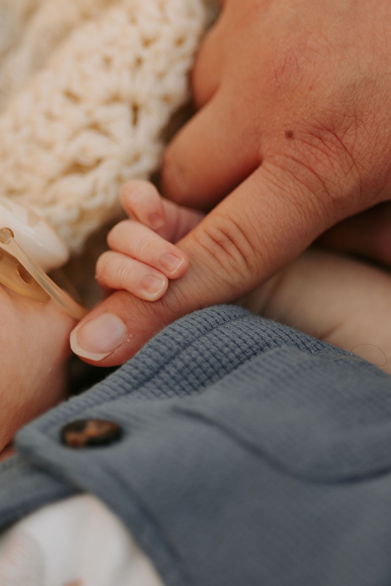A tiny baby hand gently holding an adult finger against blue fabric and white knit material.