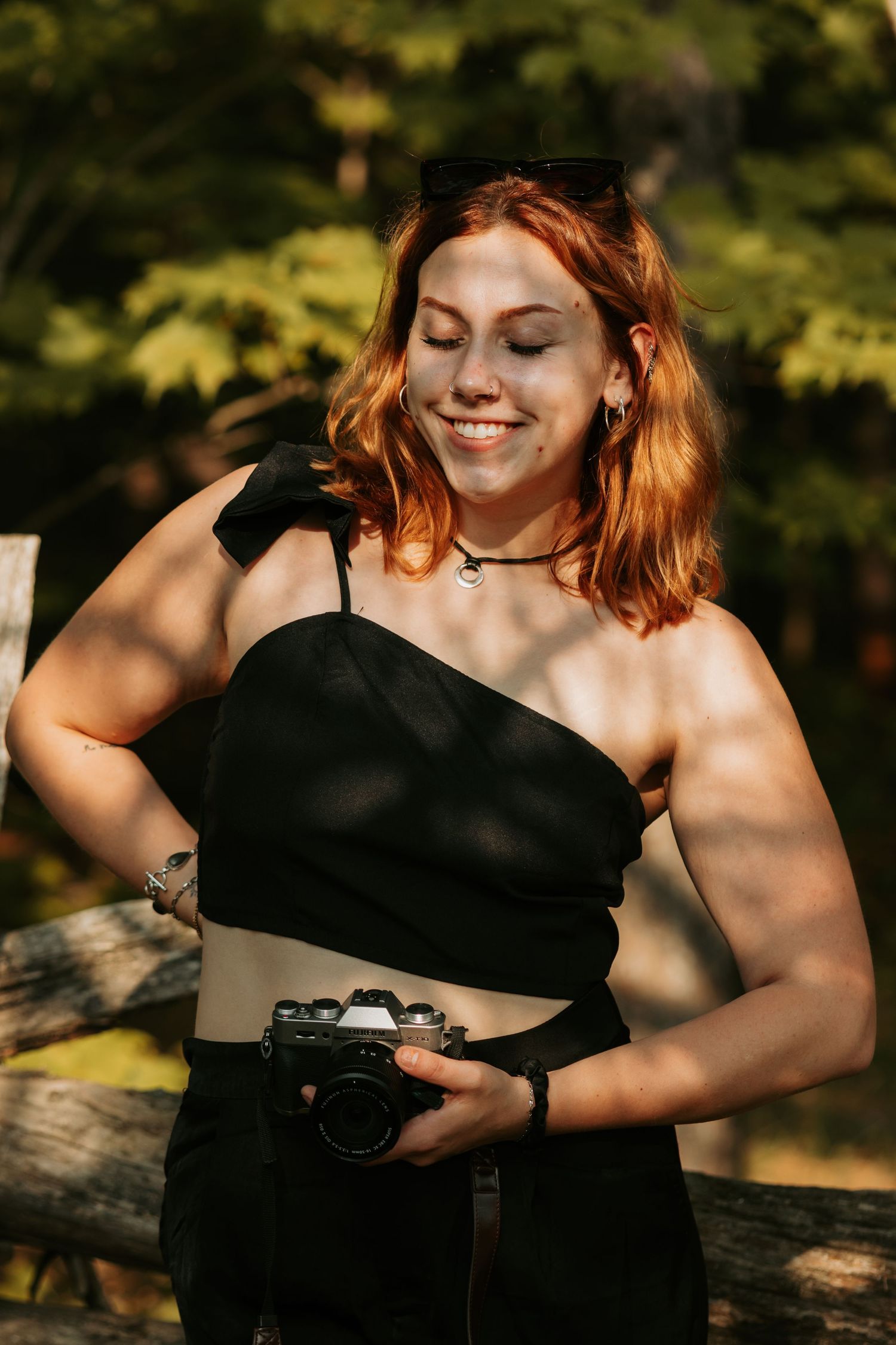 Professional photographer in a black dress holds a vintage camera in natural sunlight while smiling and posing outdoors.