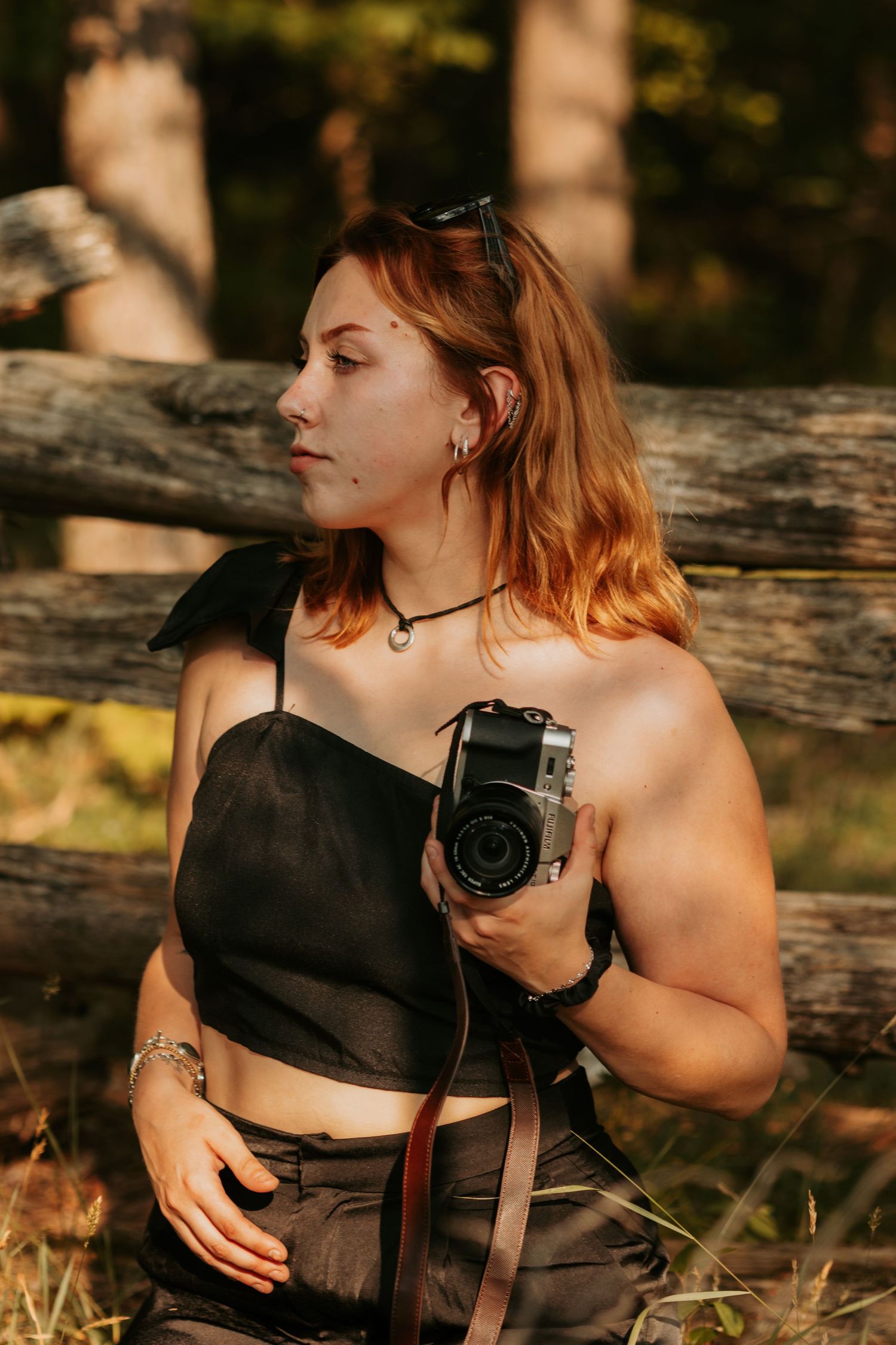 A photographer in a black crop top poses outdoors with a vintage medium format camera against rustic wooden logs.