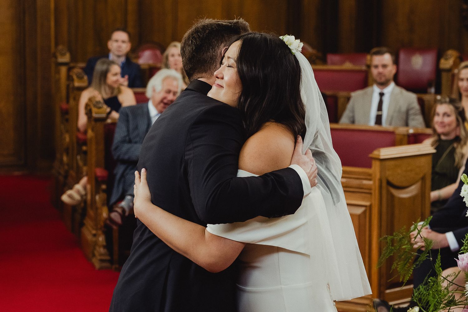 An emotional embrace between two people at a wedding ceremony in a traditional church with red carpet and wooden pews.