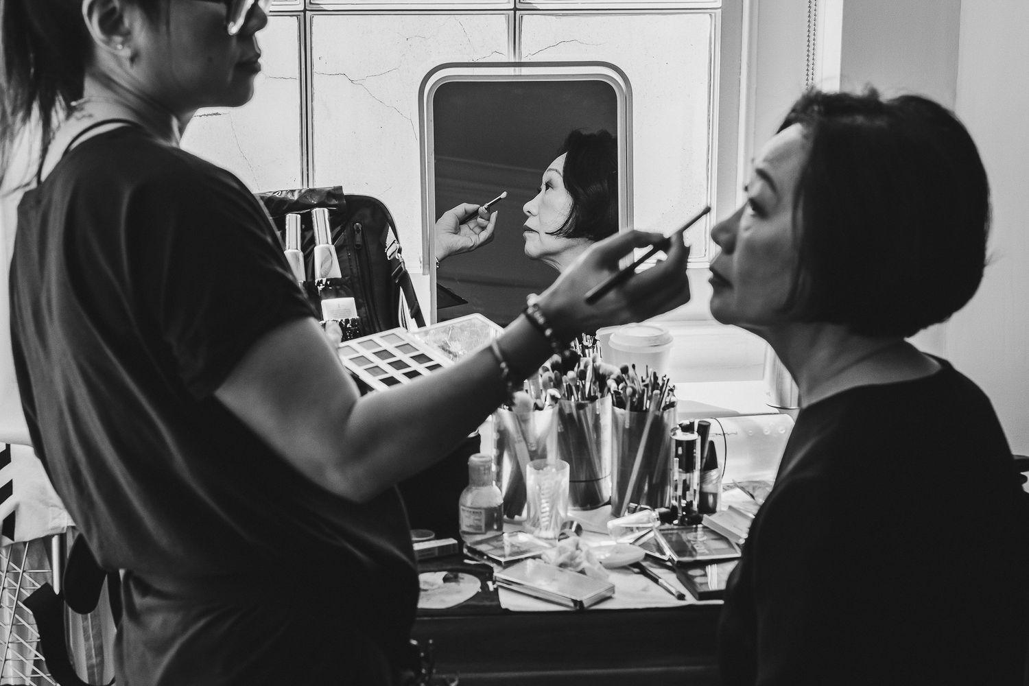 A makeup artist applies cosmetic products to someone at a vanity table in black and white photography.