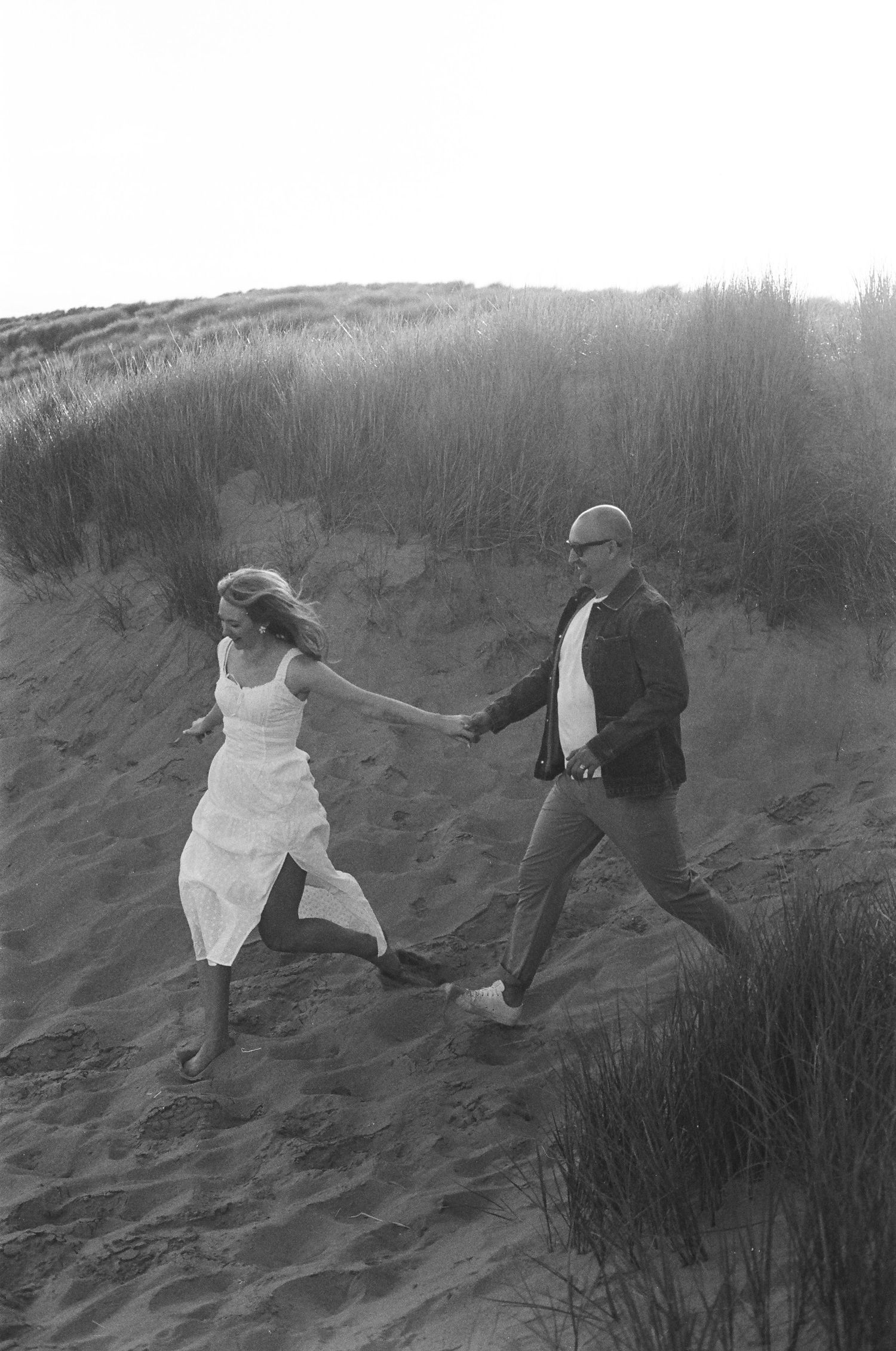 A couple in black and white holding hands and running through sand dunes on a beach.