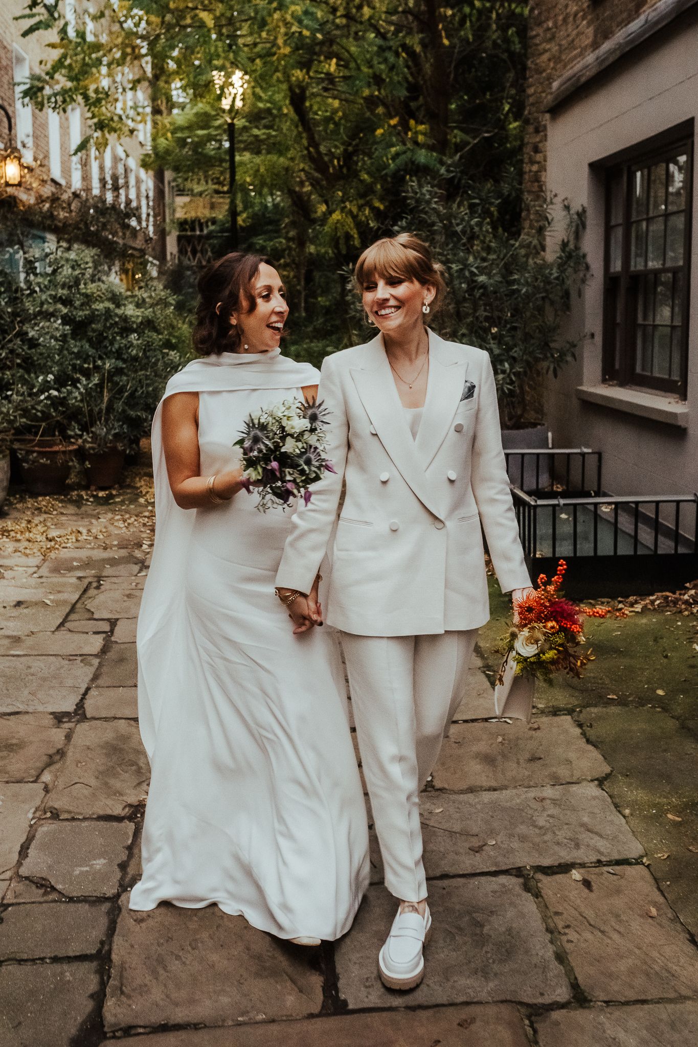 A couple in white wedding attire walk hand in hand along a stone path surrounded by autumn foliage.
