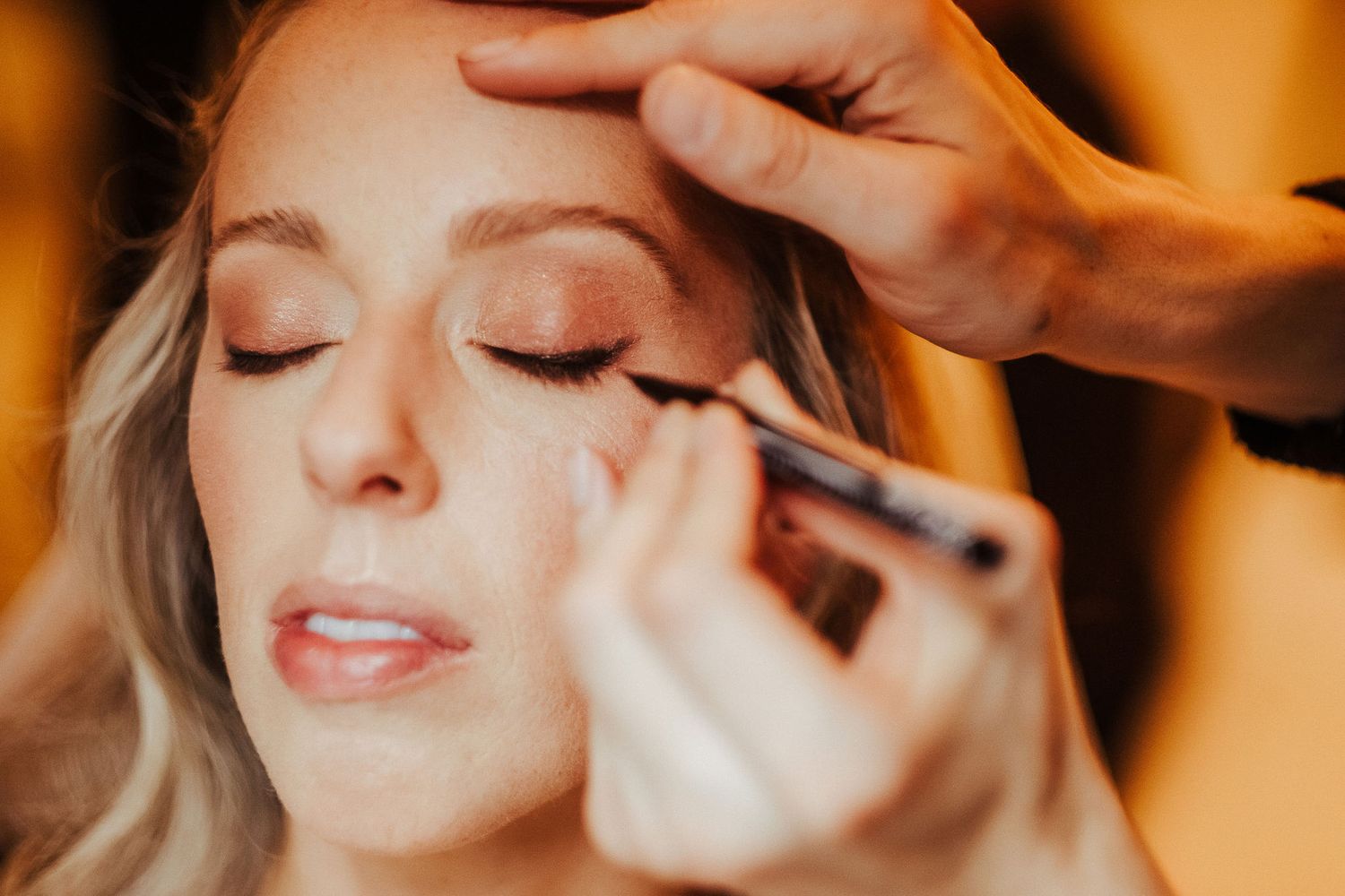 Professional makeup artist applying eye makeup on bride during wedding preparation in warm lighting.