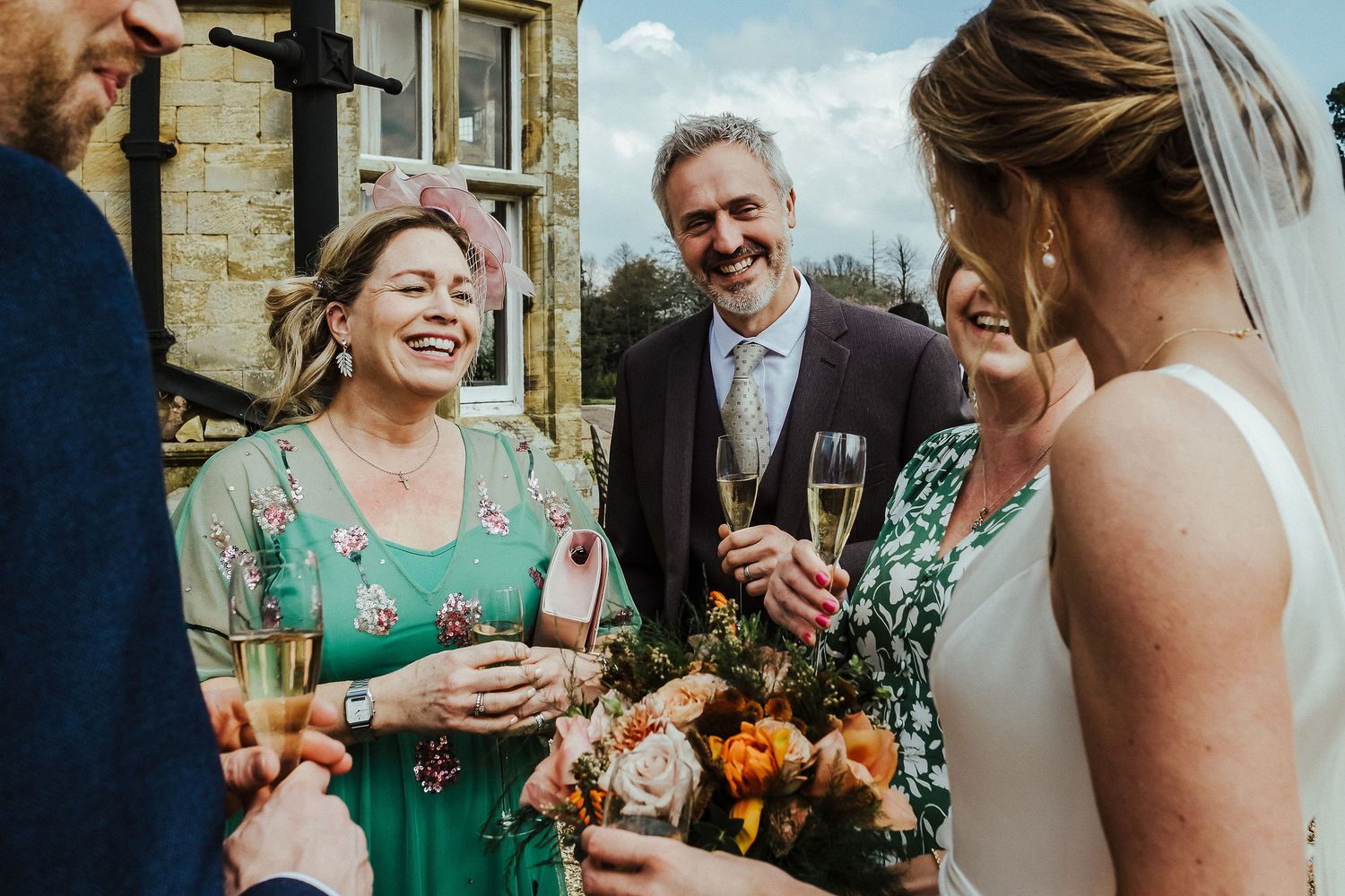 Wedding guests laugh and share champagne during outdoor reception in front of stone building.