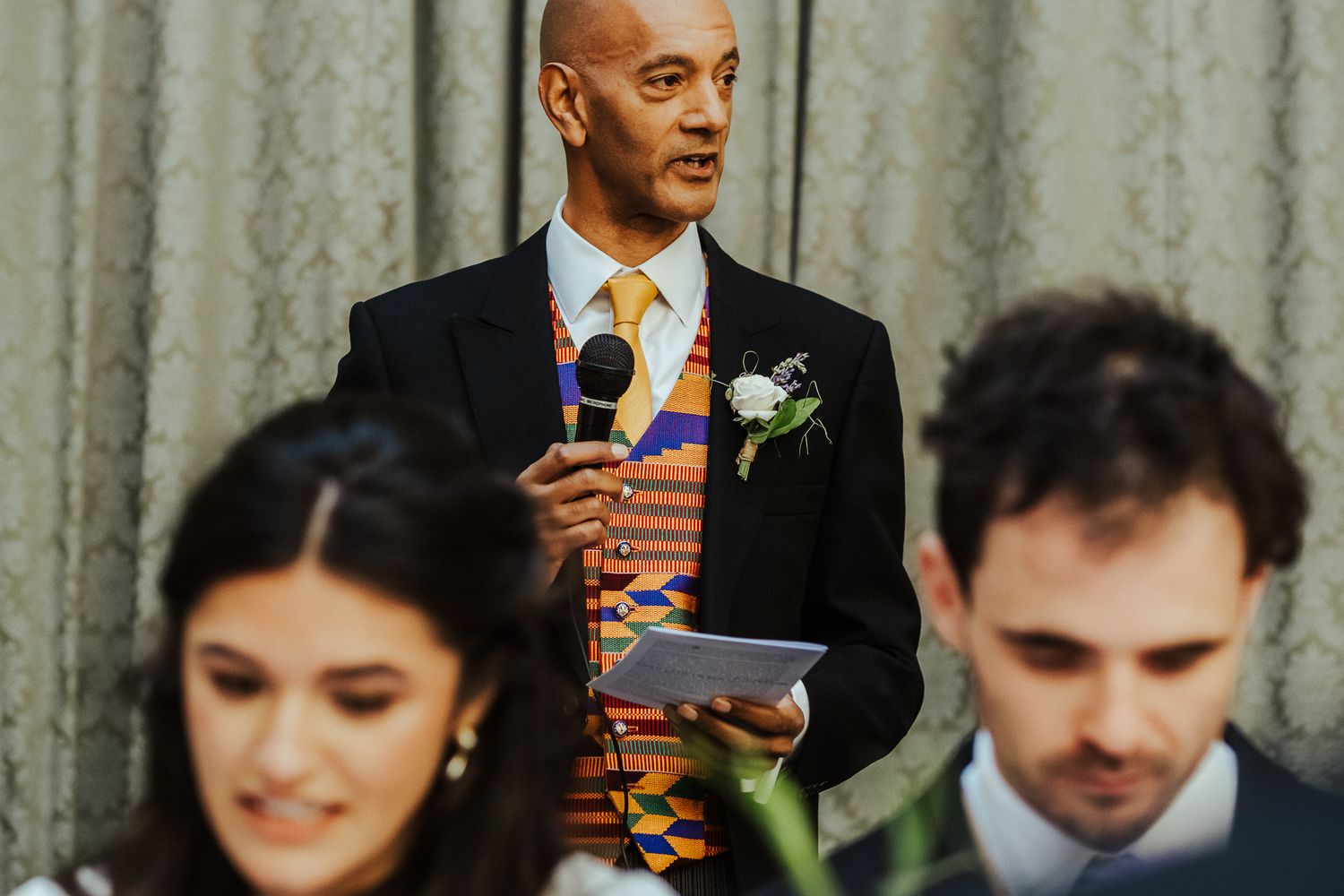 Wedding speaker delivers toast while couple listens at decorated reception table.