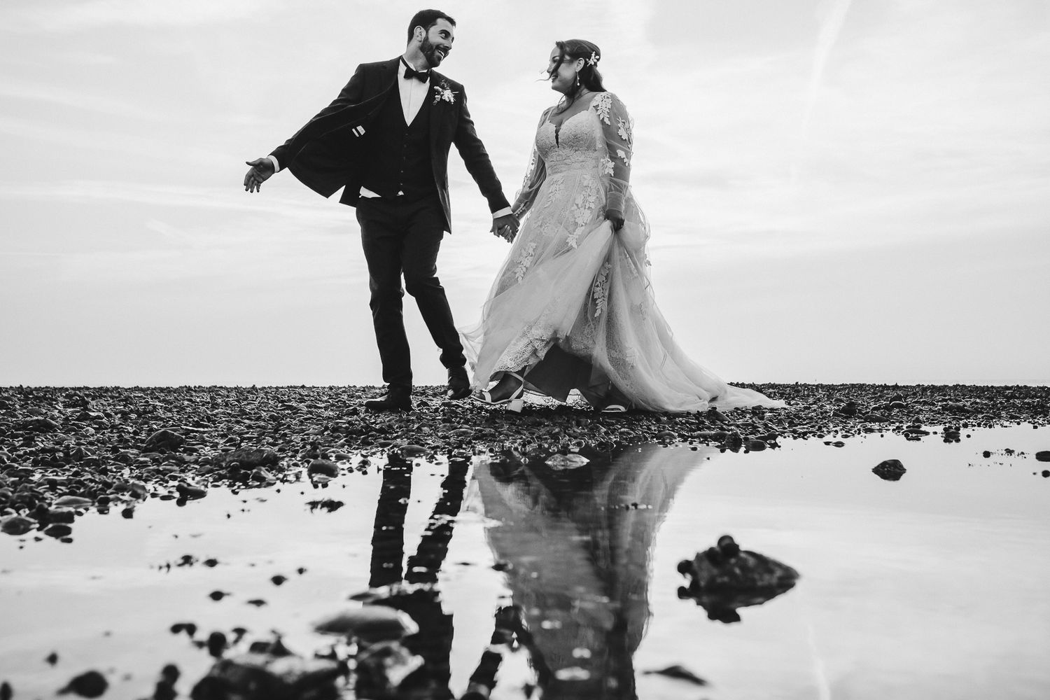 A couple in wedding attire walk hand in hand with their reflection mirrored in still water.
