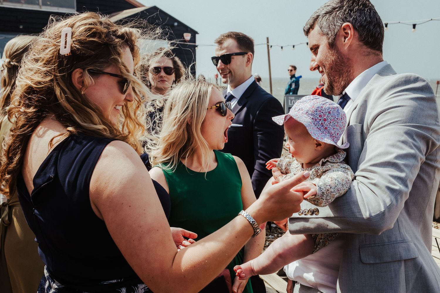 Wedding guests interact with a baby during sunny outdoor reception.