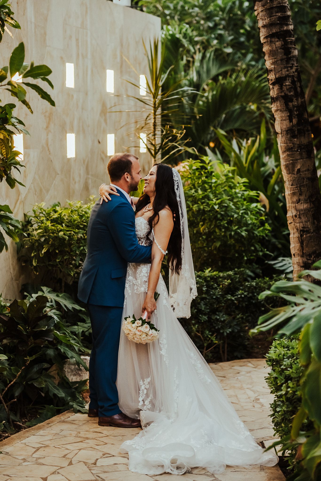 Newlyweds embrace in a lush tropical garden setting with palm trees and white walls.