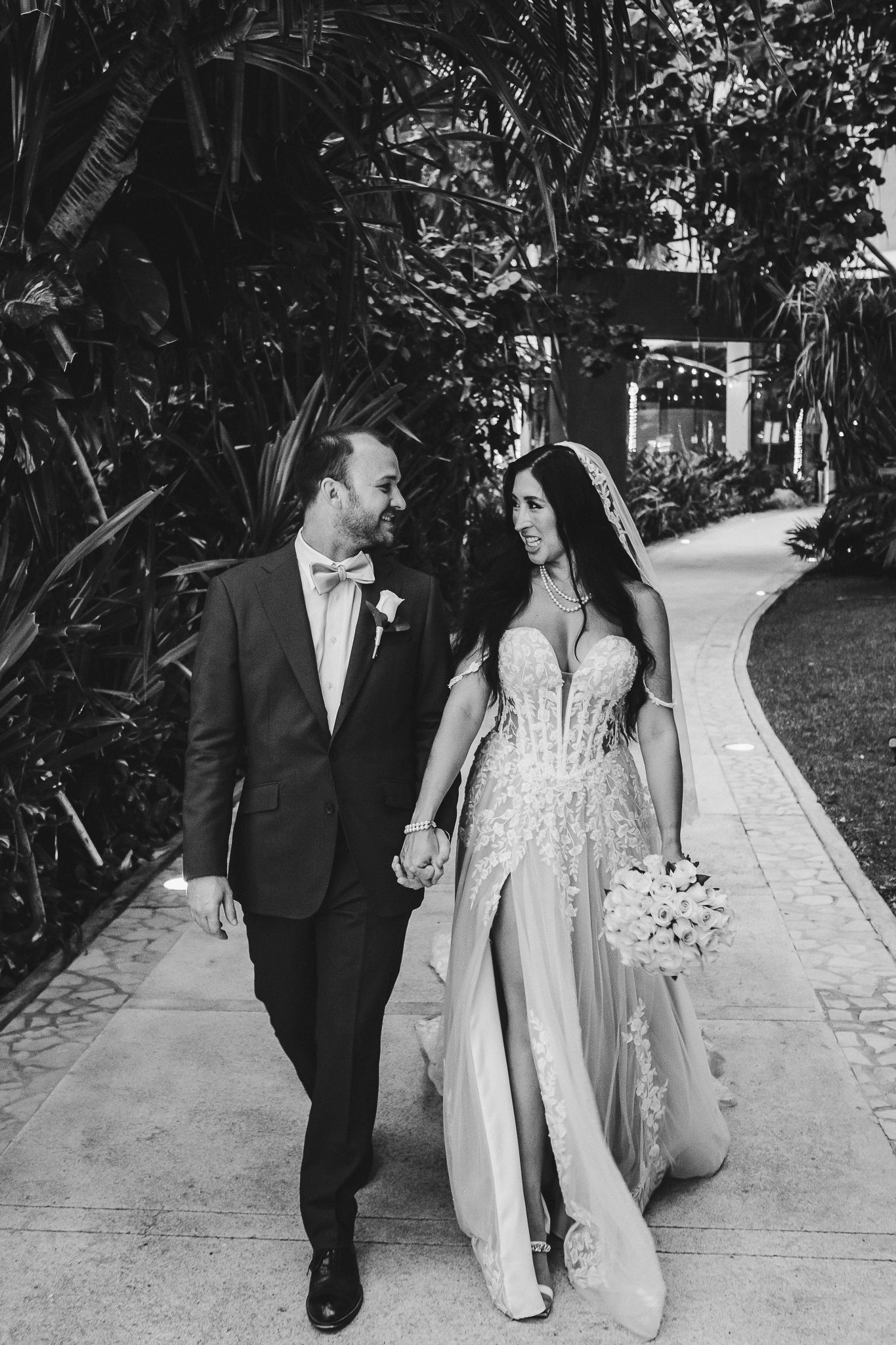 Elegant couple walking hand-in-hand down garden path in formal wedding attire.