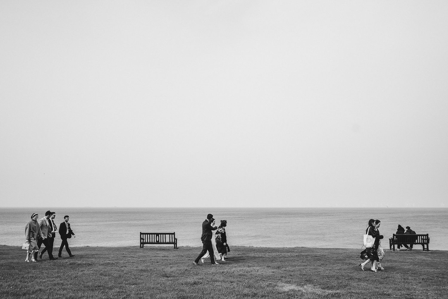 Black and white landscape of people walking along a coastal path with a bench in view.
