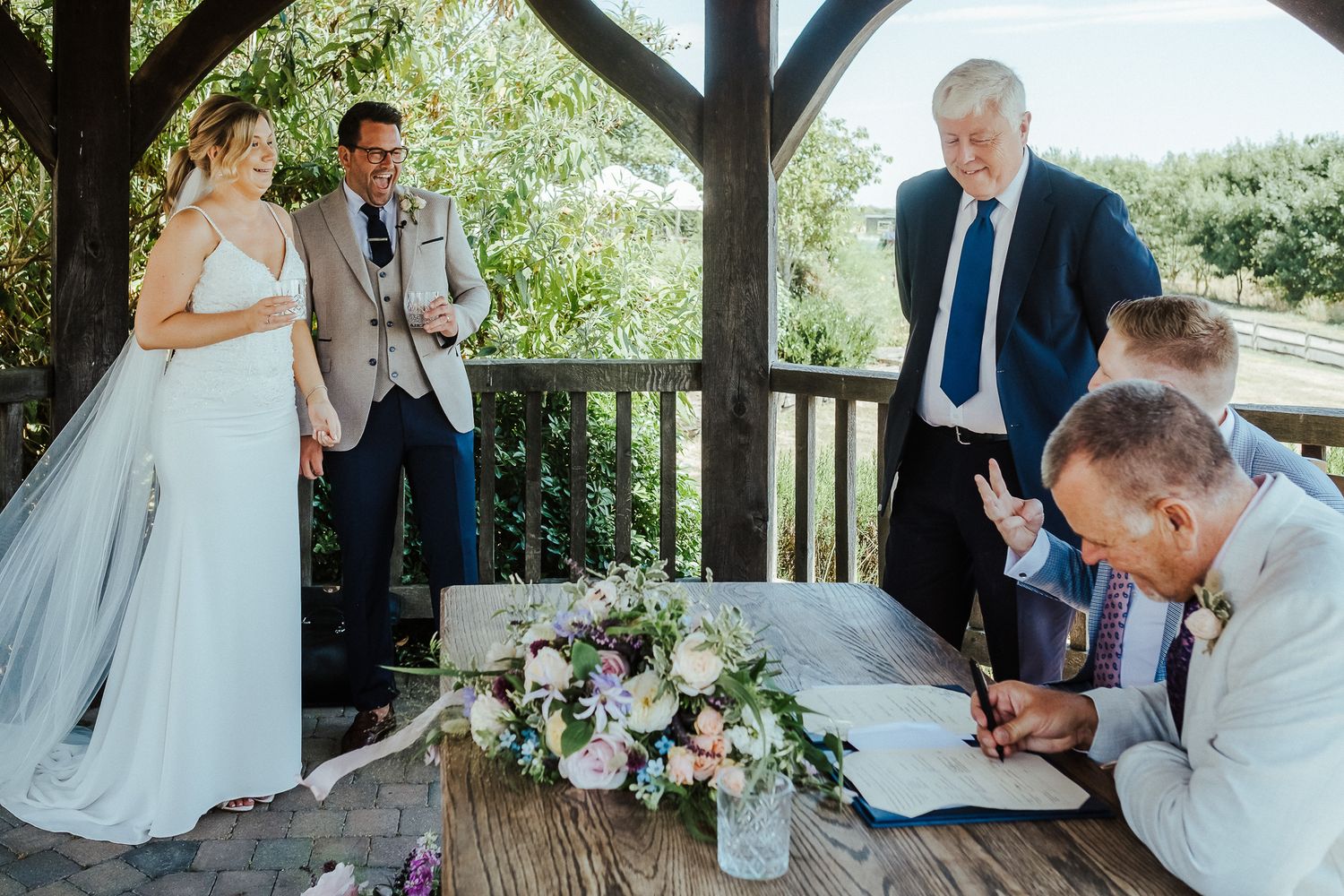 A couple signing marriage documents under a wooden gazebo decorated with floral arrangements.