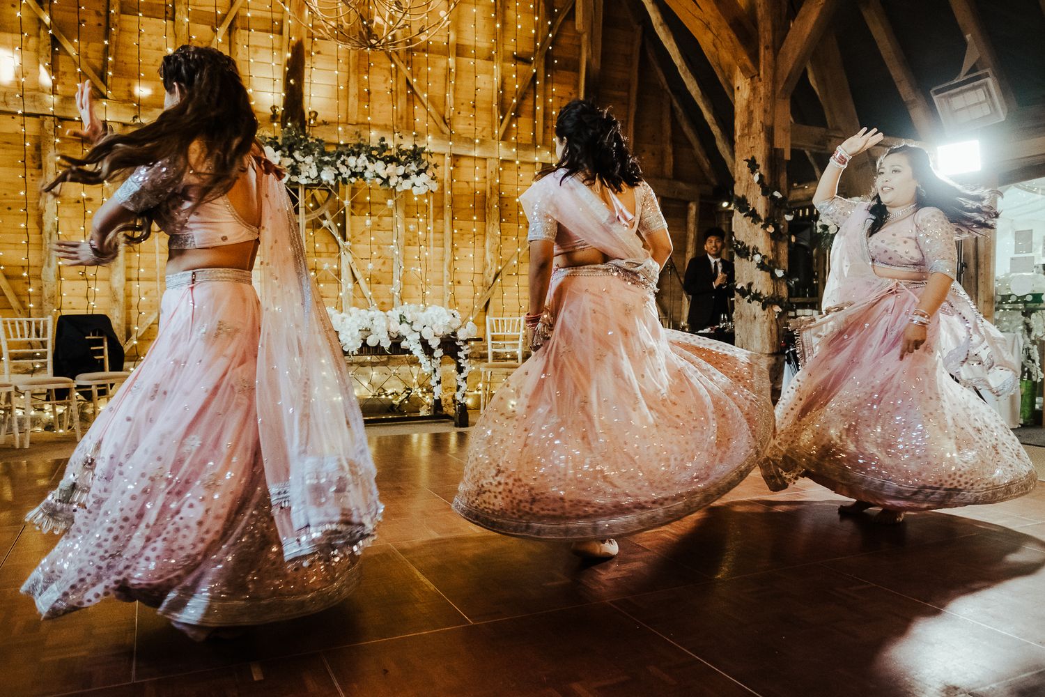 Three dancers in pink lehengas twirl on a dance floor under warm lighting.