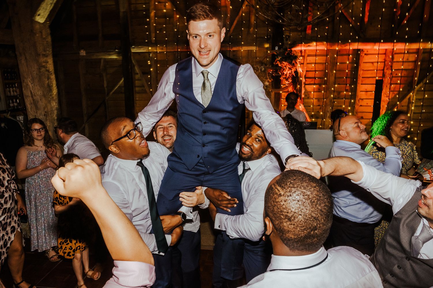 Wedding party dancing energetically at dimly lit reception with string lights.