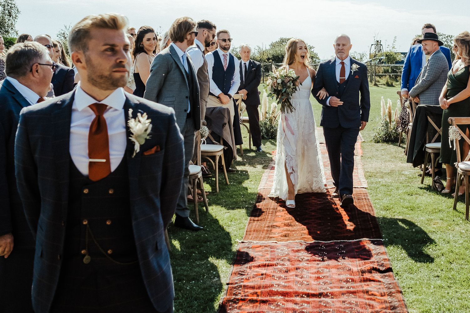 An outdoor wedding ceremony with vintage rugs lining the aisle and guests gathered on a sunny day.