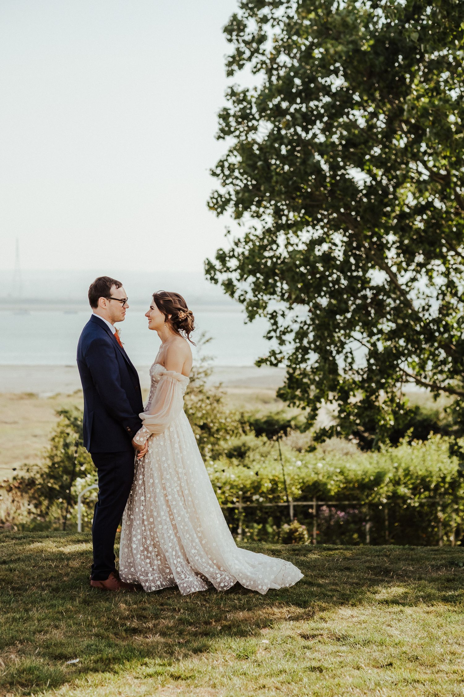 A couple in wedding attire stands on a hilltop overlooking water, surrounded by lush greenery at golden hour.
