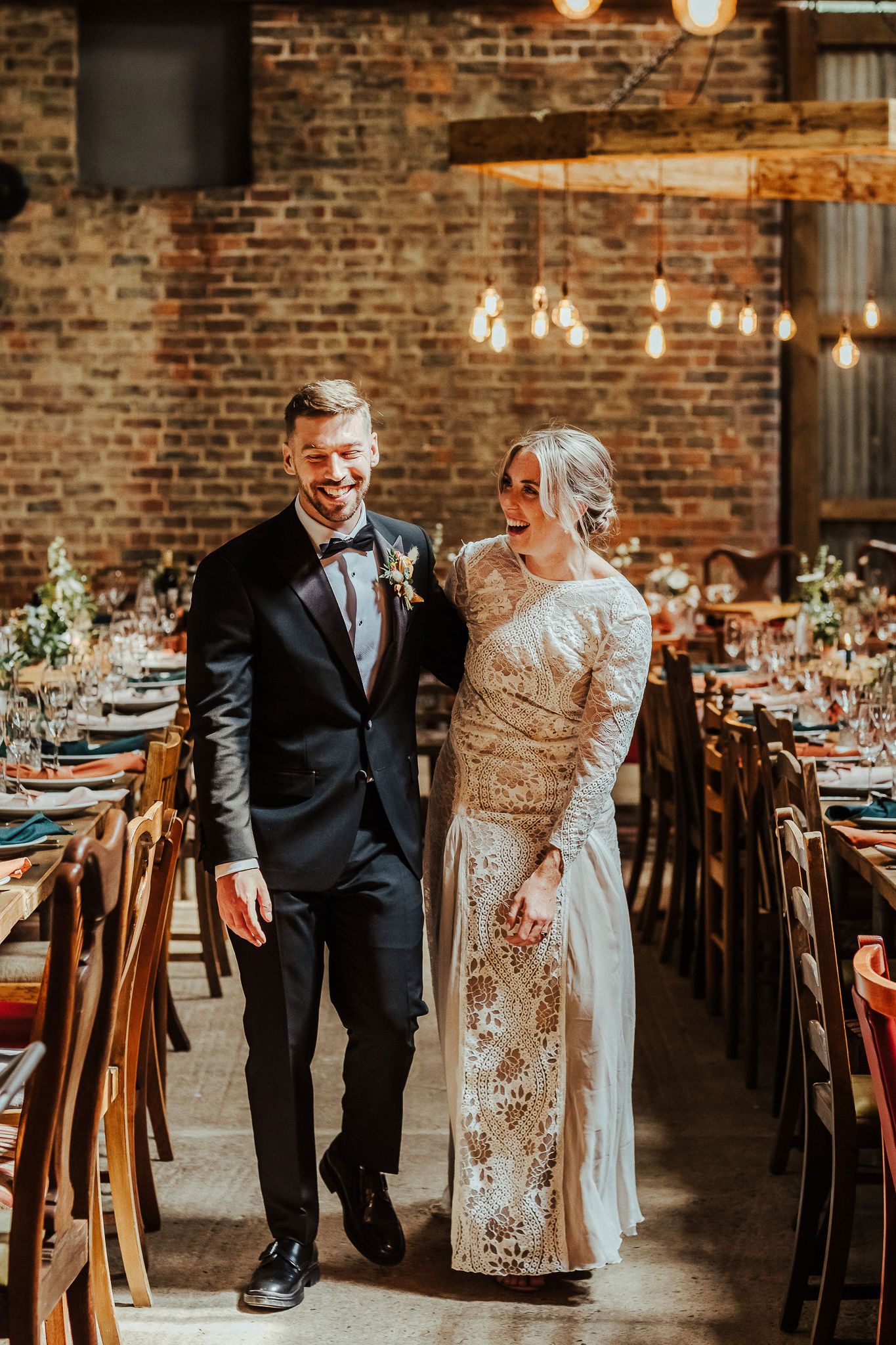 Wedding party walking through rustic brick venue with elegant table settings and string lights.
