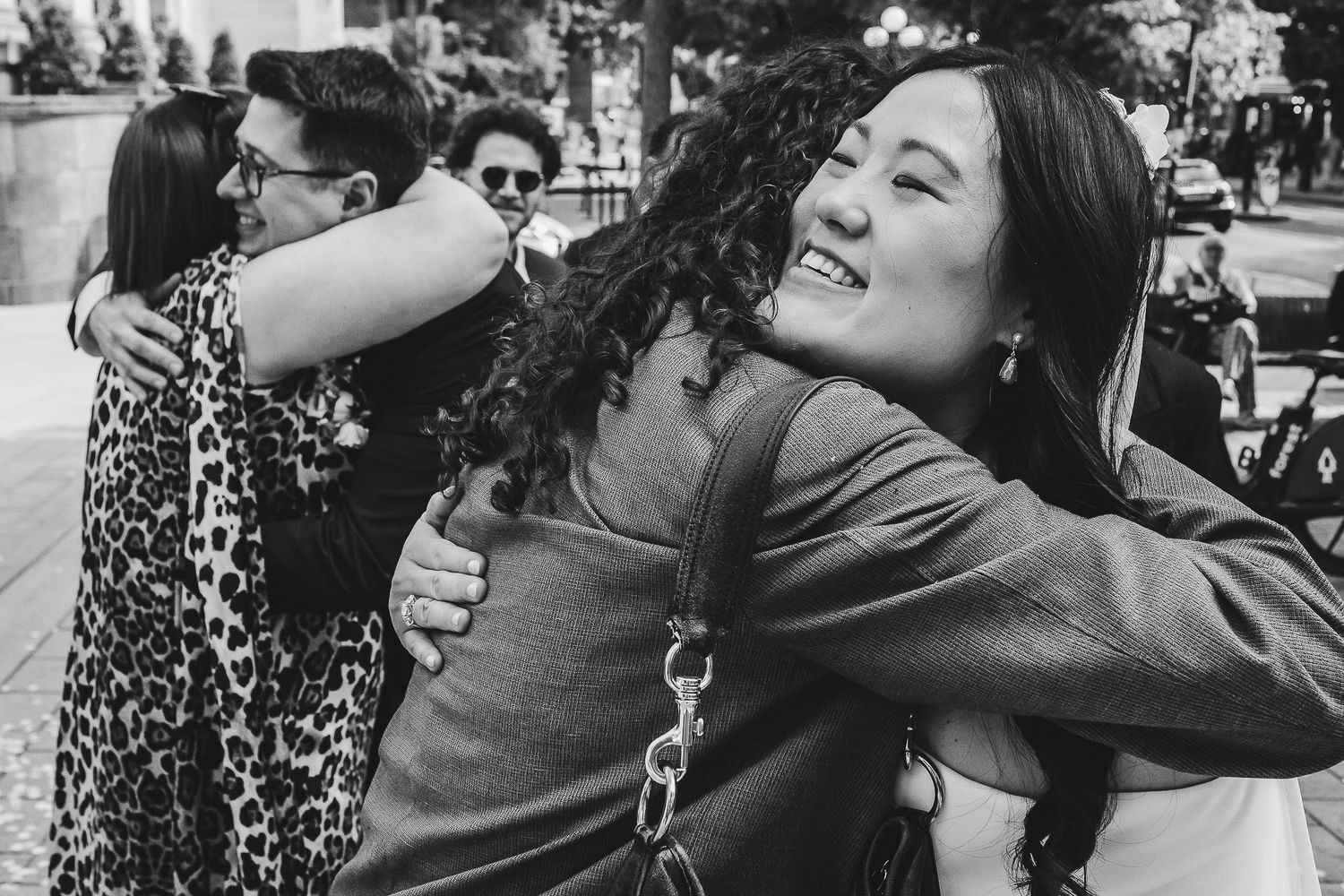 Black and white photo showing two people sharing a joyful embrace while others gather in the background.