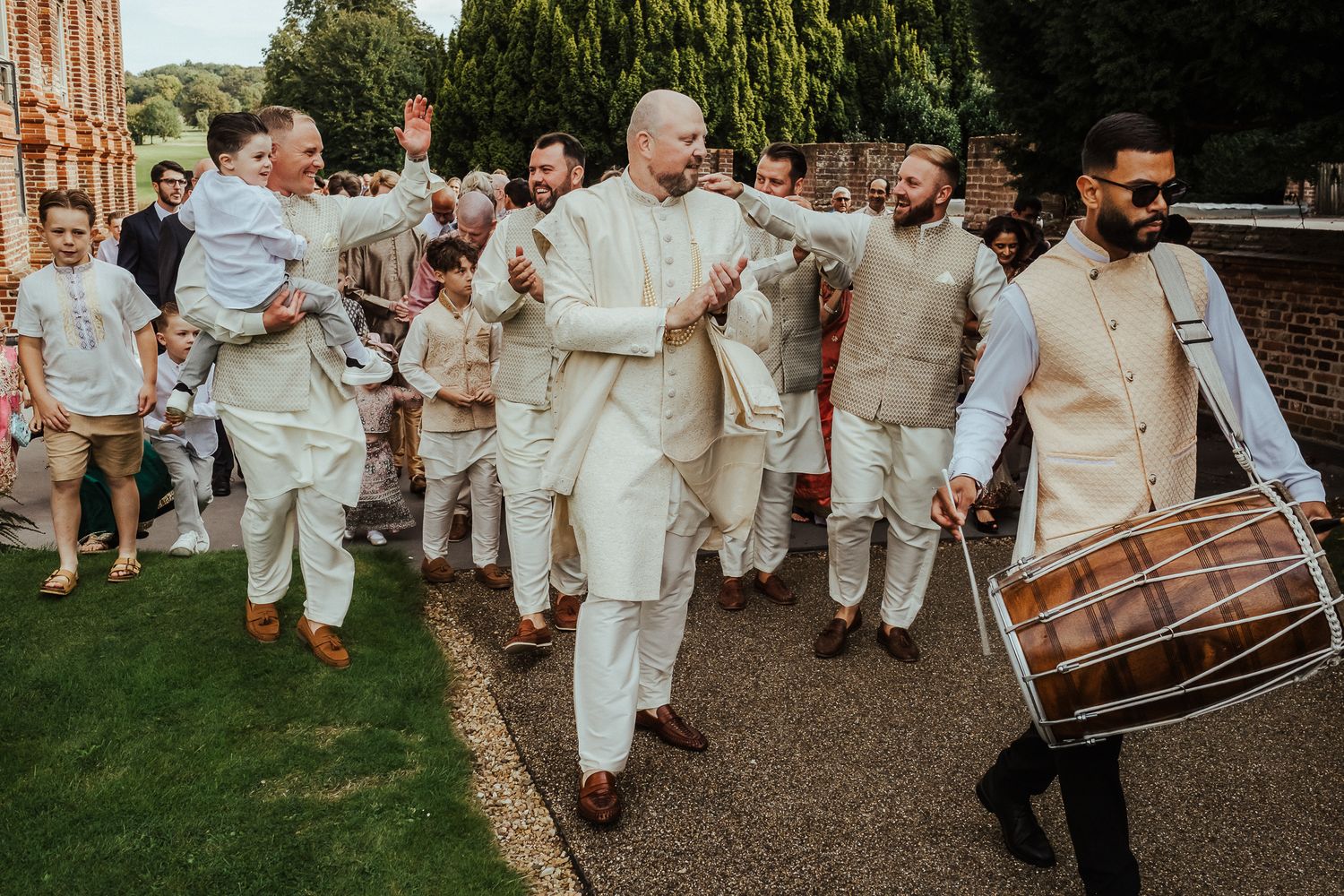 A joyful wedding procession with dhol drummer and guests walking along a garden path in natural linen outfits.