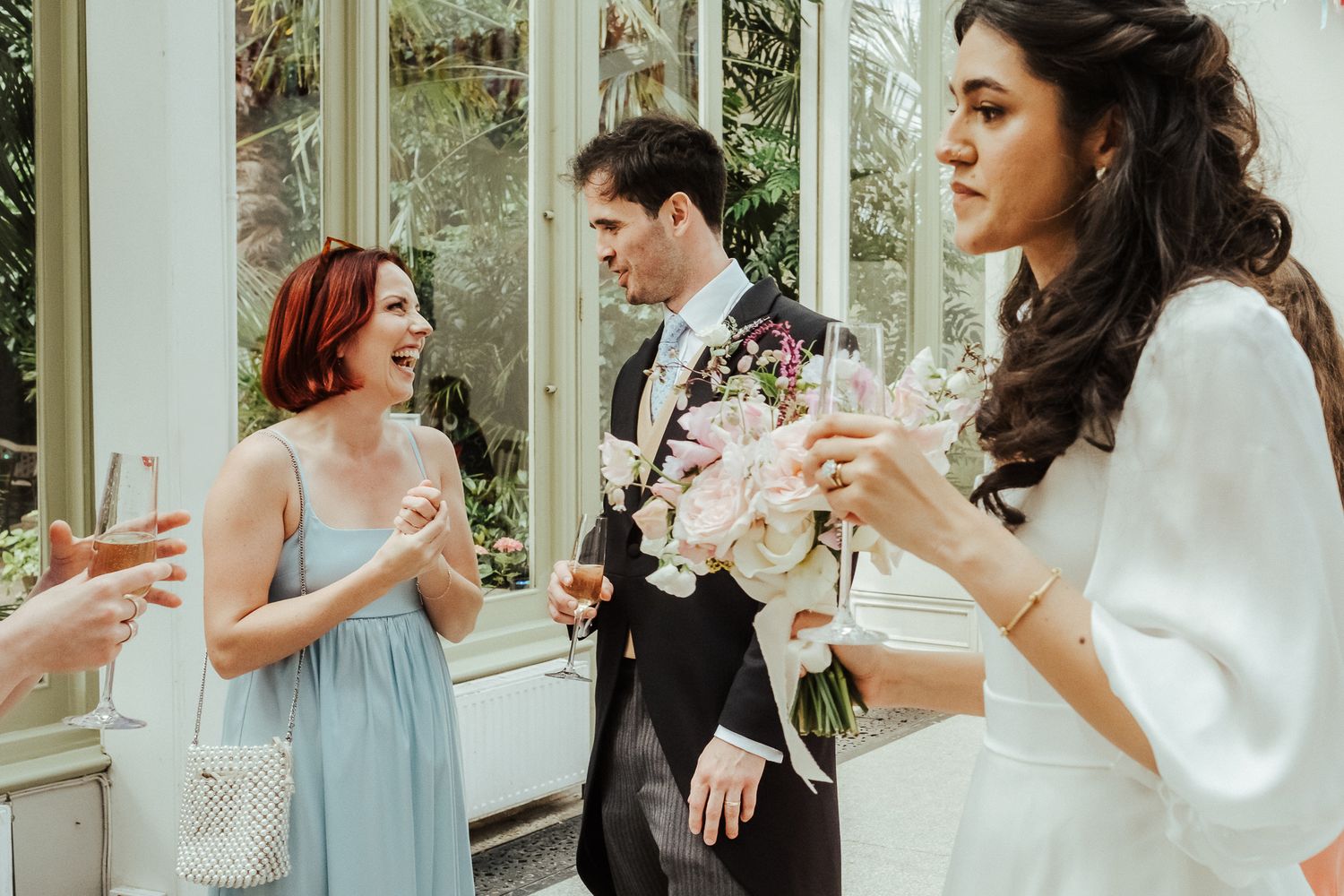 Wedding party members chatting in conservatory with floral bouquets.