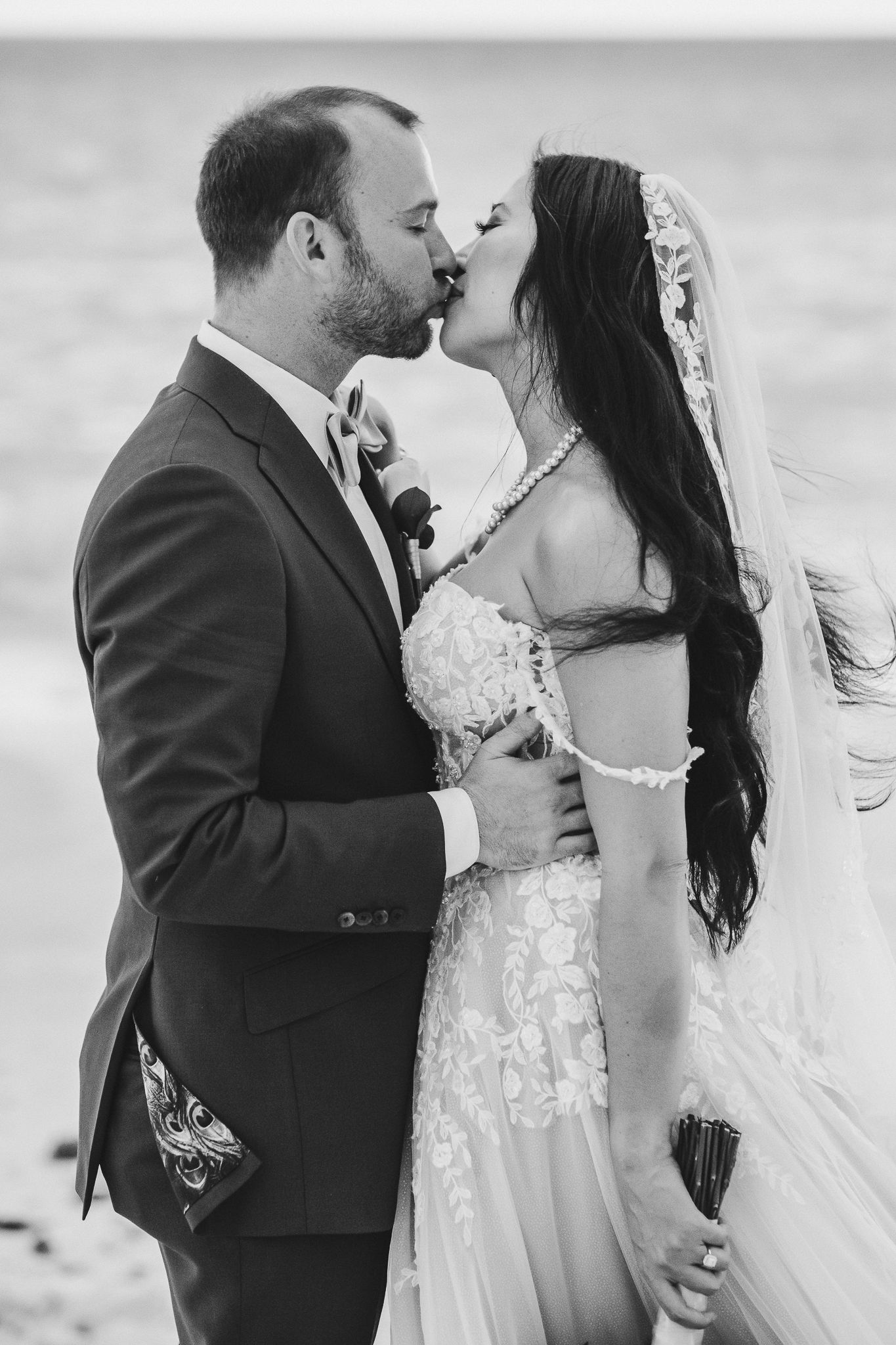 A romantic black and white beach wedding photo of newlyweds sharing their first kiss in formal attire.