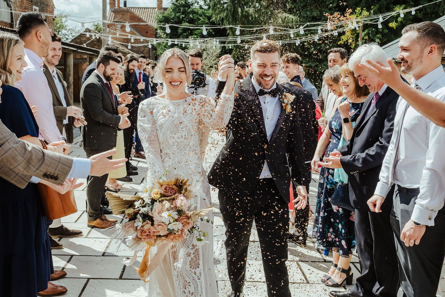 Guests shower the couple with confetti during an outdoor celebration under string lights.