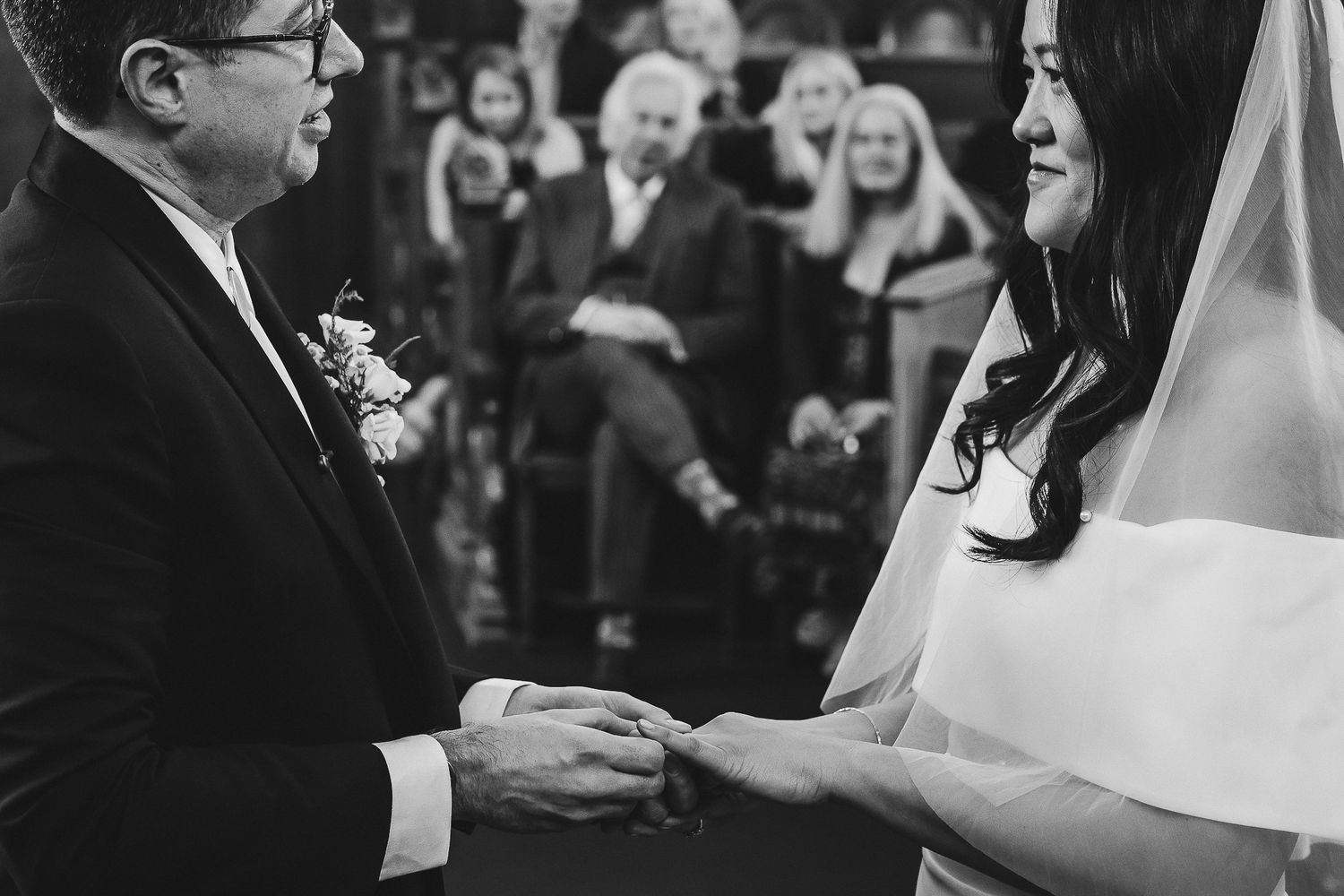 Black and white photo of a wedding ceremony ring exchange with guests watching in background.