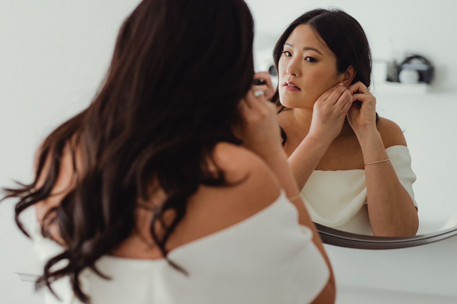 Woman wearing white tank top putting on earrings while looking in mirror during morning routine.