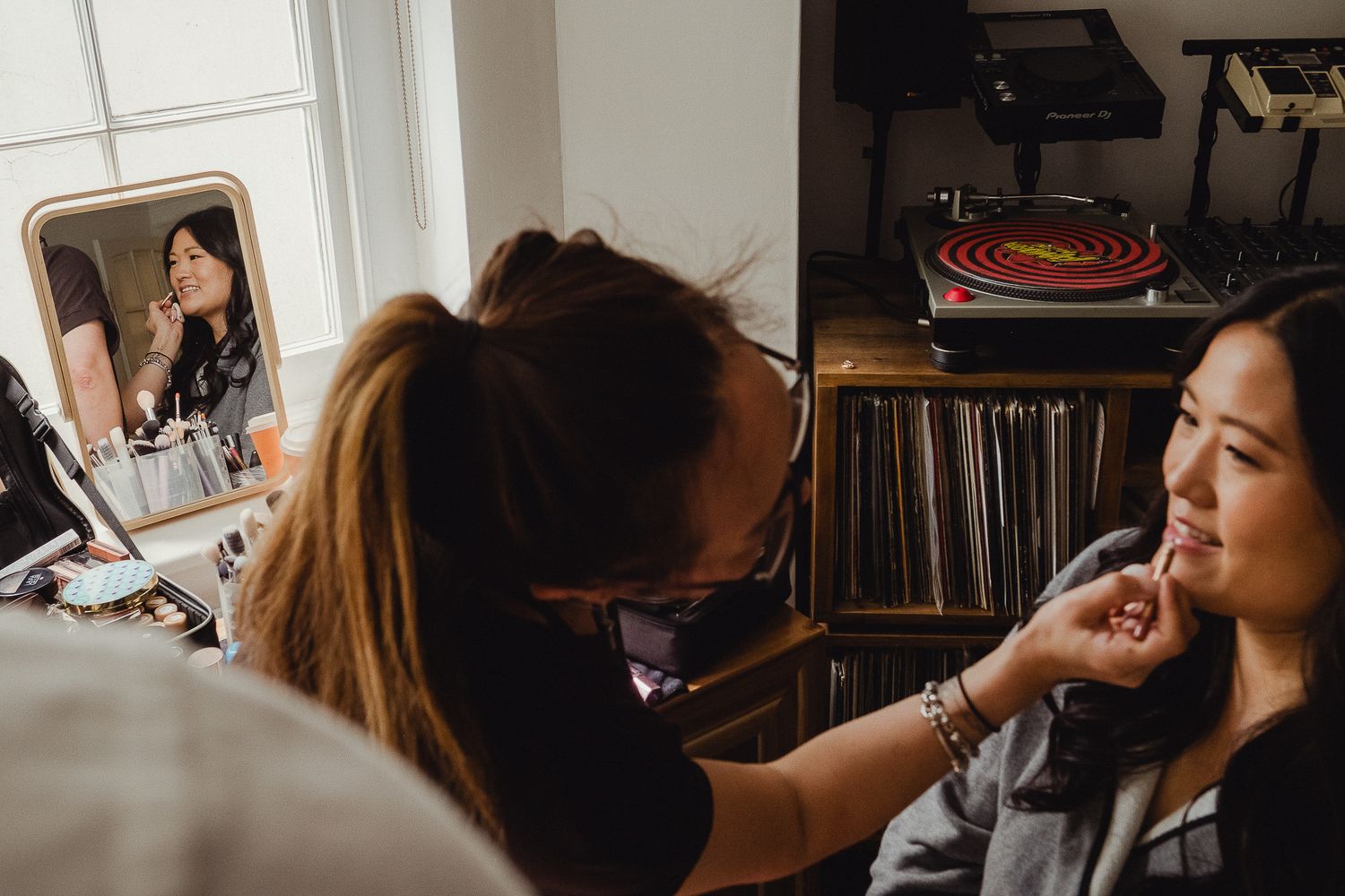 Makeup artist applies cosmetics on client while working next to vinyl record collection in warm lighting.